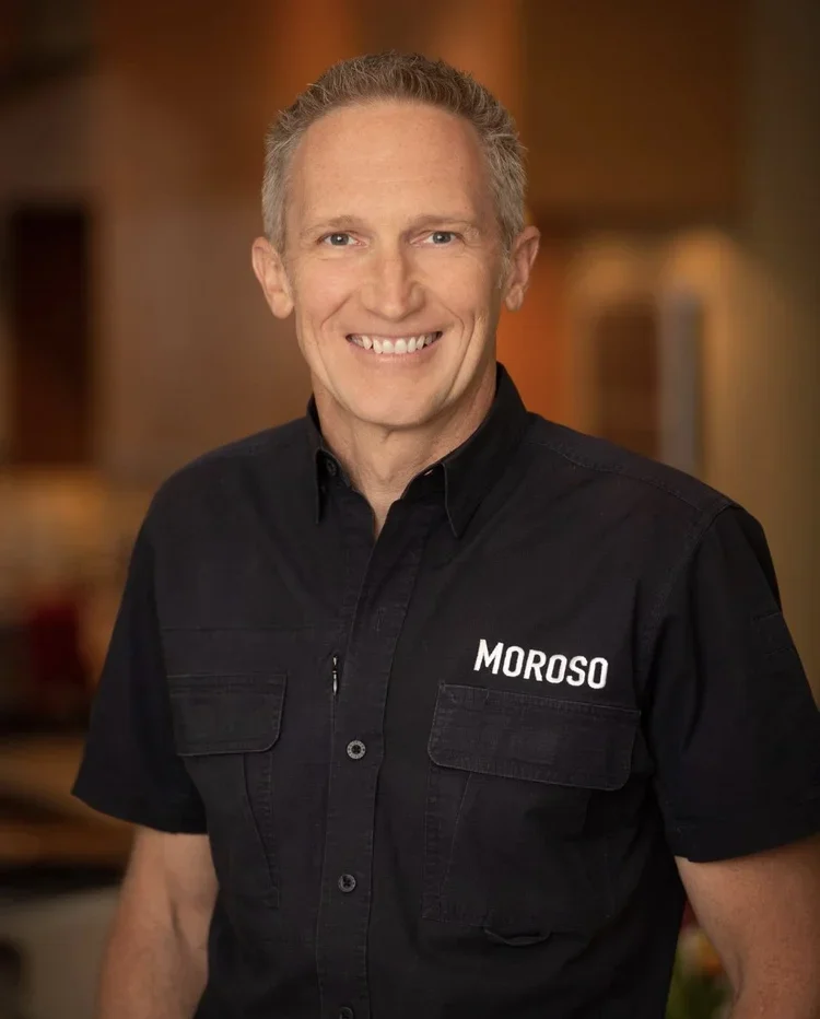 A smiling man with short gray hair wearing a black short-sleeved button-up shirt with the word 'MOROSO' embroidered on the left chest. The background is blurred warm-toned indoor setting.