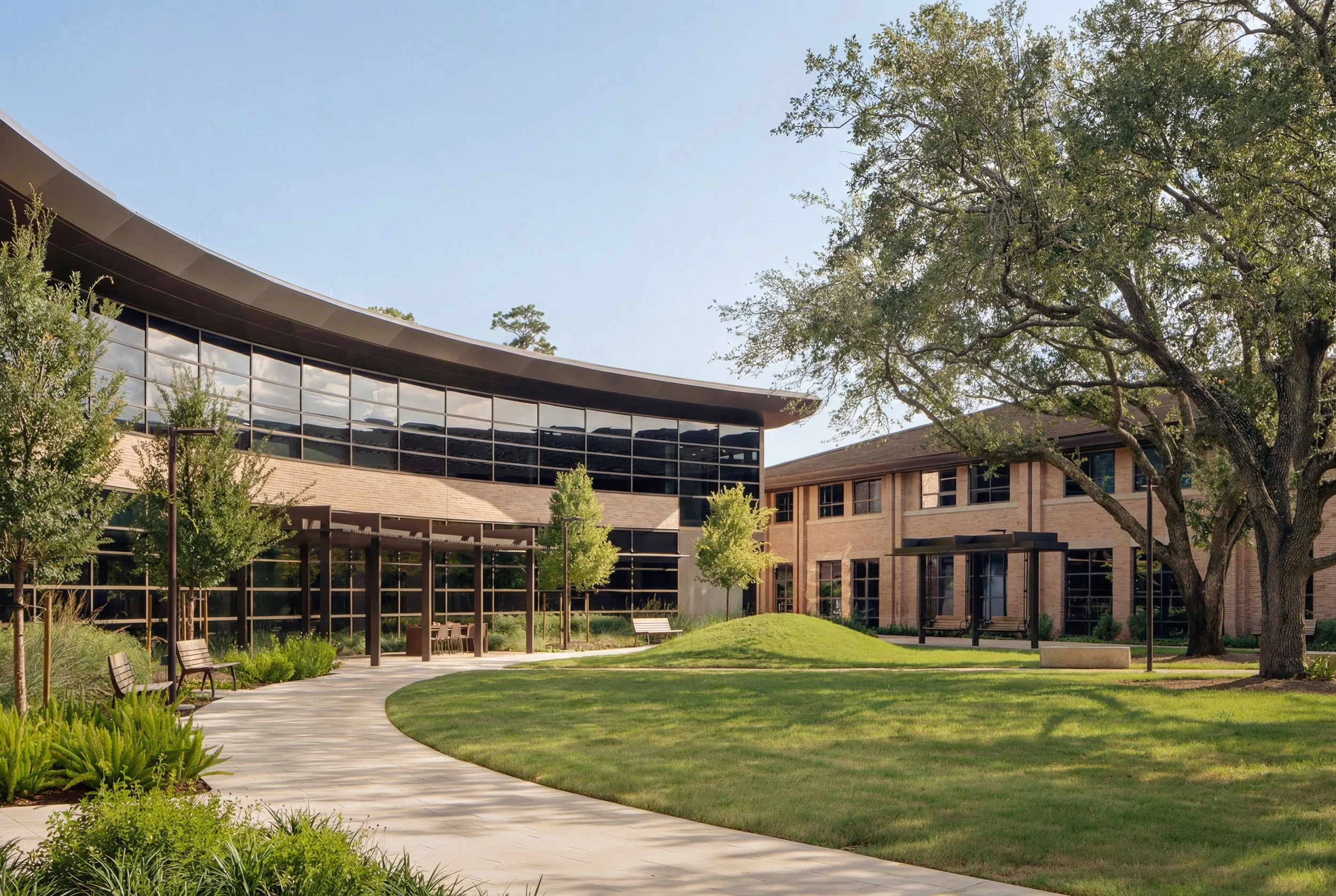 Modern building with curved glass windows, surrounded by a landscaped green lawn with trees, benches, and a pathway.