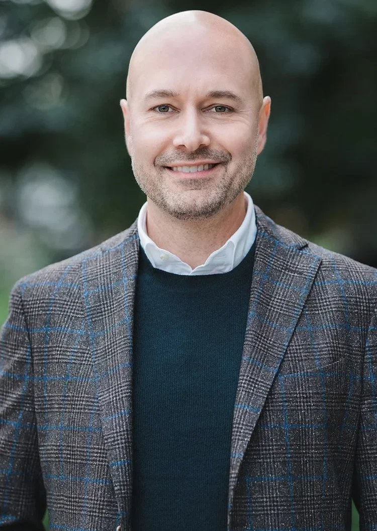 A smiling bald man in a plaid blazer, white shirt, and navy sweater standing outdoors with a blurred green background.
