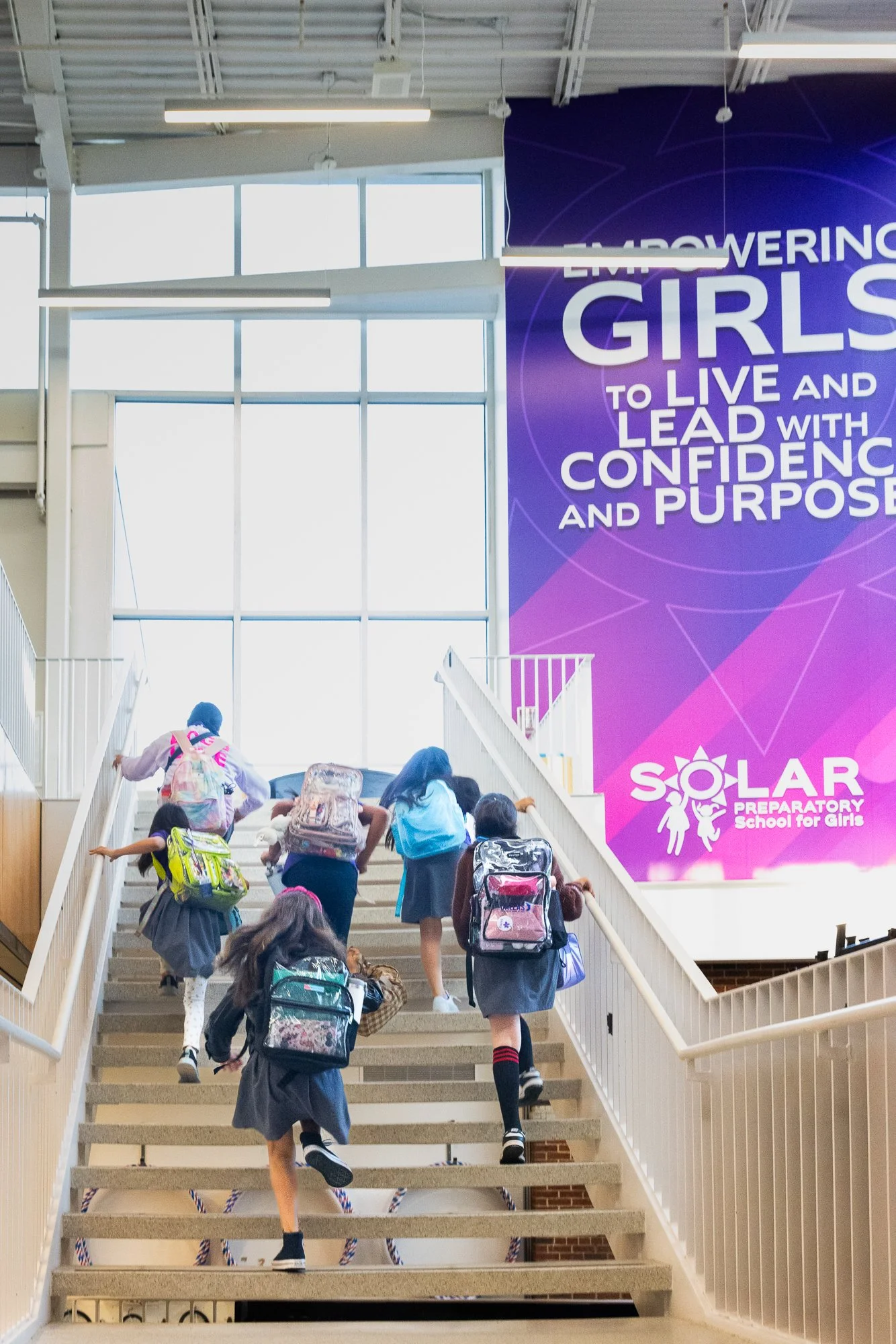 Students with backpacks climbing stairs inside a school building with a large purple banner encouraging empowering girls to live and lead with confidence and purpose, and a logo for Solar Preparatory School for Girls.