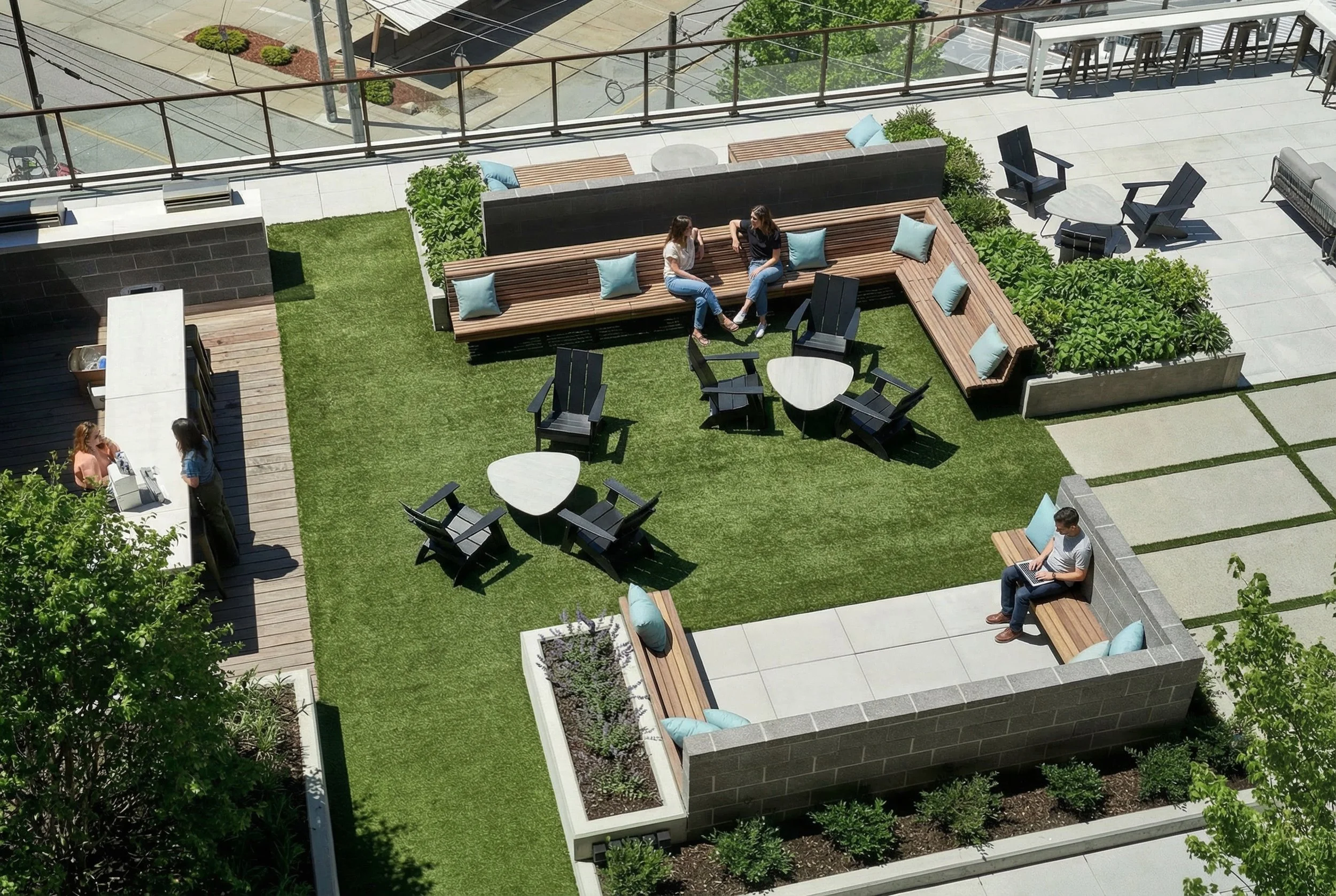 A rooftop outdoor lounge area with wooden benches, black chairs, and small tables. Four people are sitting, talking, or working on laptops, and a barista is serving coffee to two women at a counter. There are landscaped plants and artificial grass.