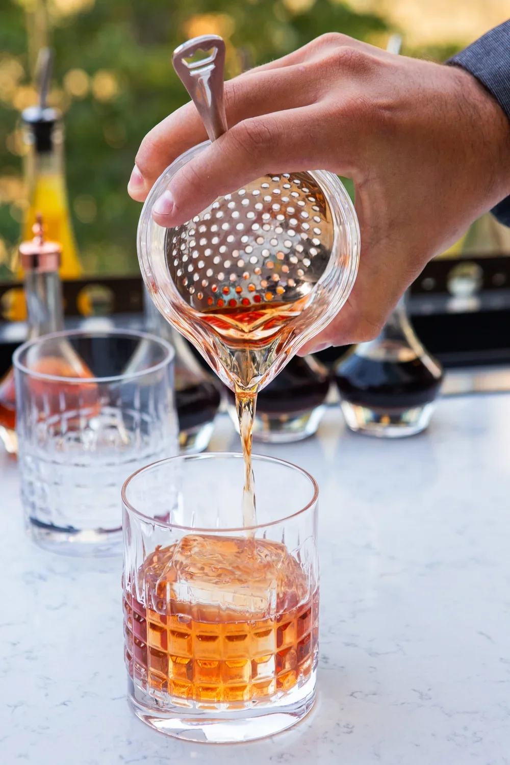 A person pours rosé wine through a metal strainer into a glass with ice on a marble surface, with other glasses and bottles of wine in the background.