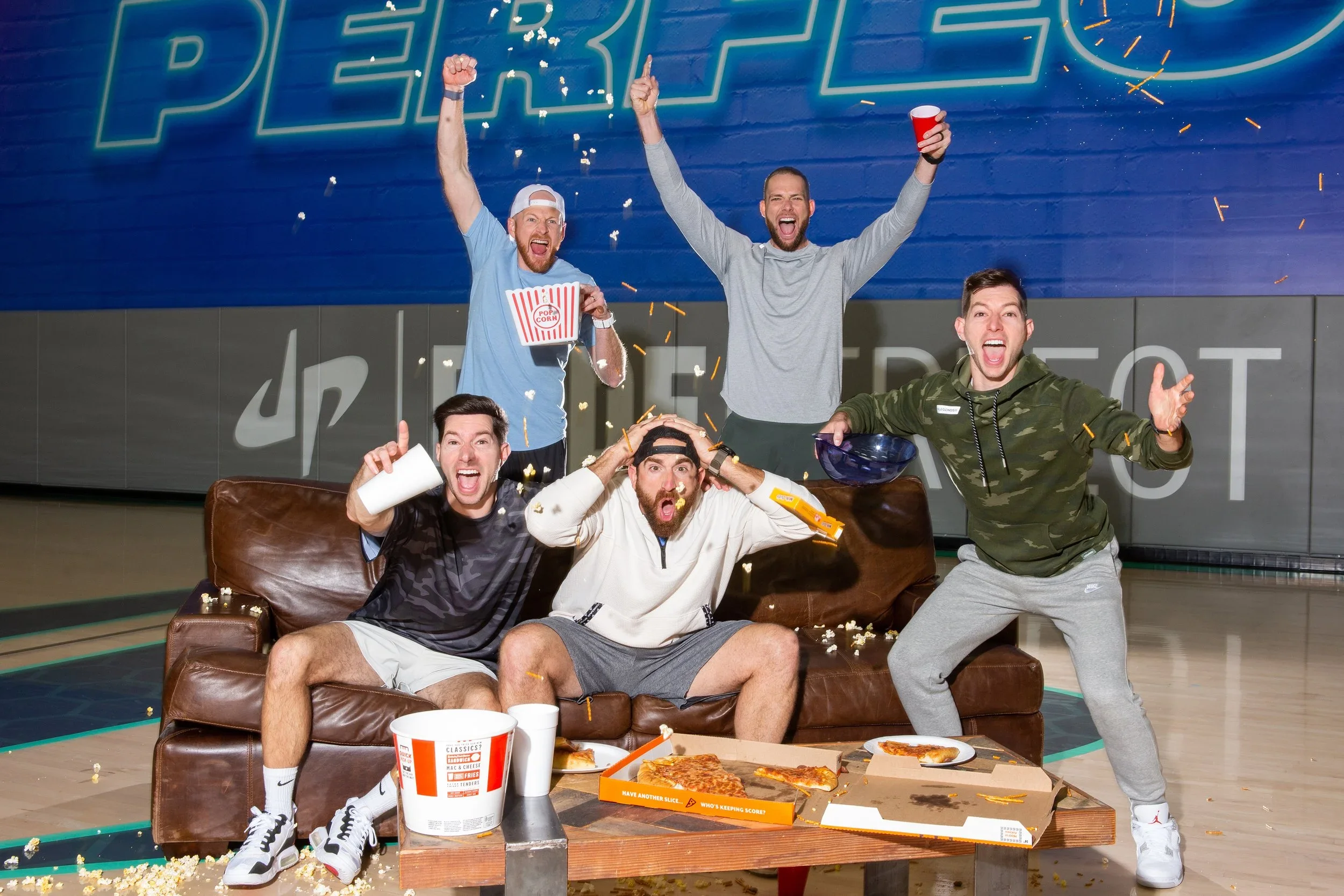 Group of five young men celebrating after playing bowling, sitting and standing on a couch in a bowling alley, with pizza, popcorn, drinks, and popcorn box on table and couch, some of them cheering and making expressive faces, colorful background wit