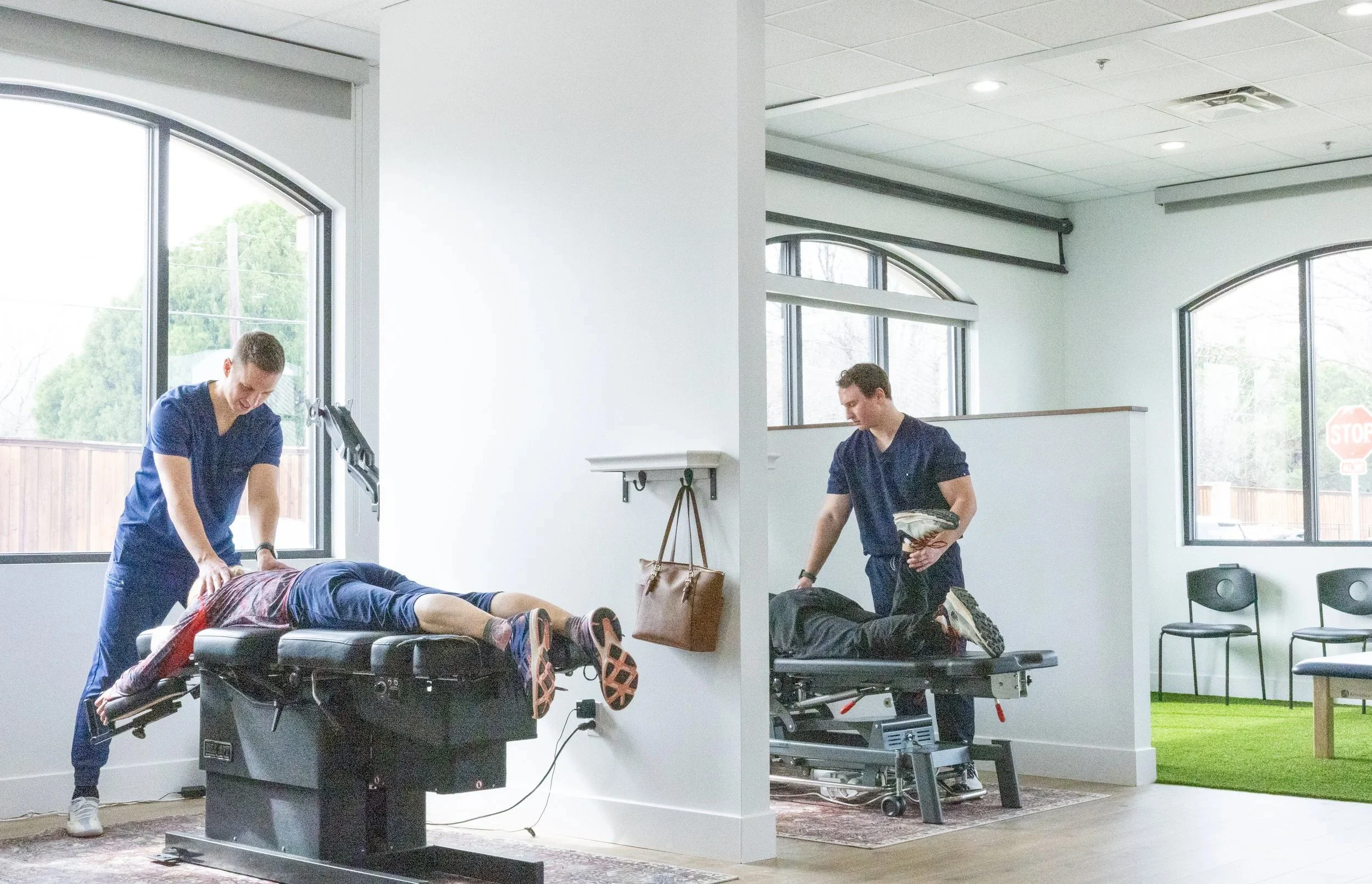 Two physical therapists working with patients on chiropractic adjustments in a bright, modern clinic divided by a wall.