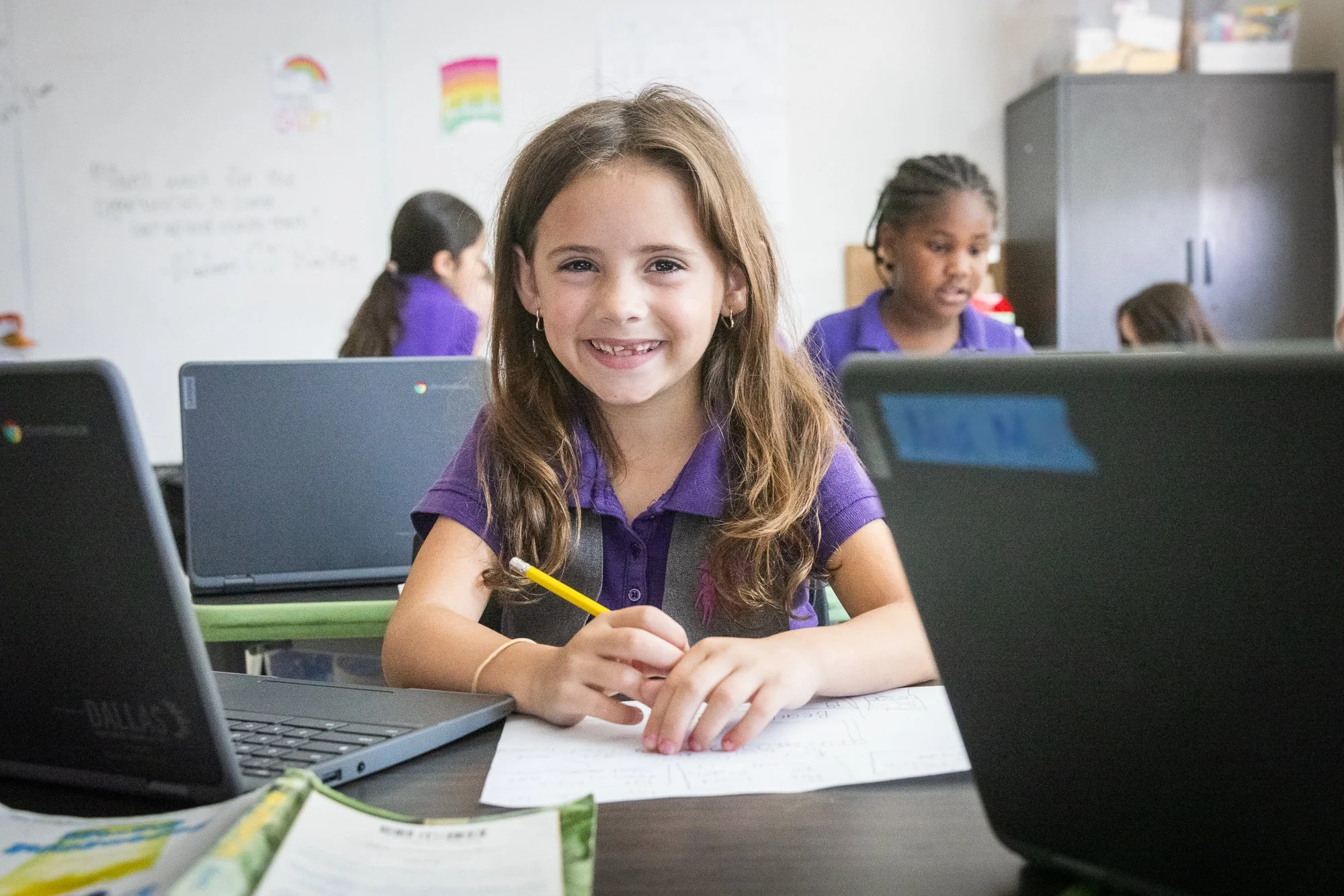 A young girl smiling at a classroom desk with laptops, holding a yellow pencil, with other students working in the background.