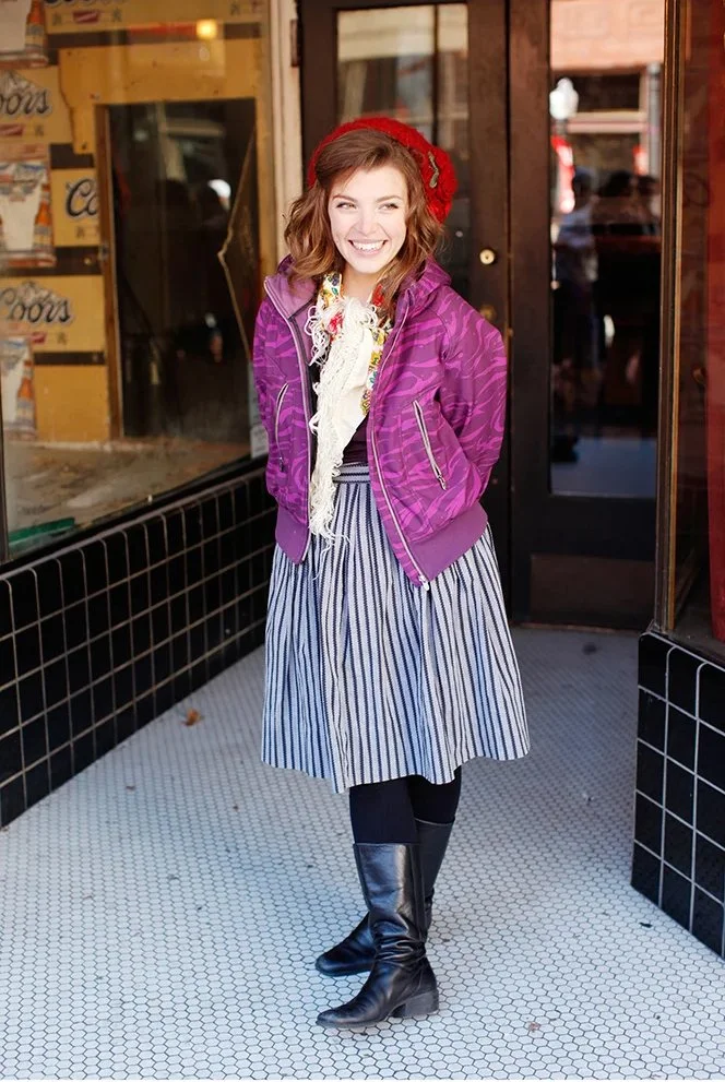 A young woman standing outside a shop, smiling and looking at the camera, wearing a red beret, purple jacket, striped skirt, and black knee-high boots.