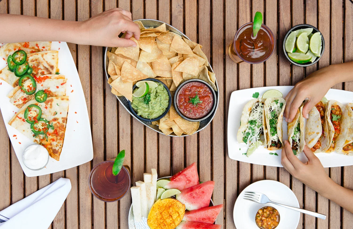 Top-down view of a wooden table with various Mexican dishes including slices of pizza, chicken tacos with cilantro and lime, a plate of watermelon slices, a bowl of guacamole, salsa, tortilla chips, and drinks with lime wedges.