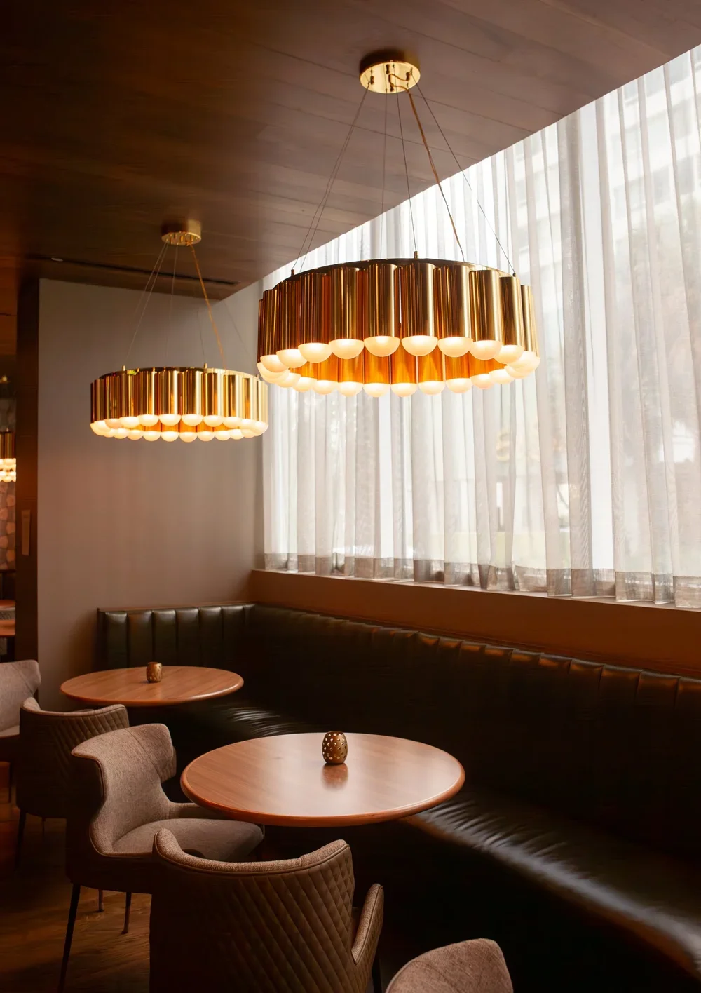 Interior of a modern restaurant or lounge with two large gold and white hanging pendant lights, wooden ceiling, and sheer curtains covering windows. There are round wooden tables, cushioned chairs, and a leather banquette along the wall.