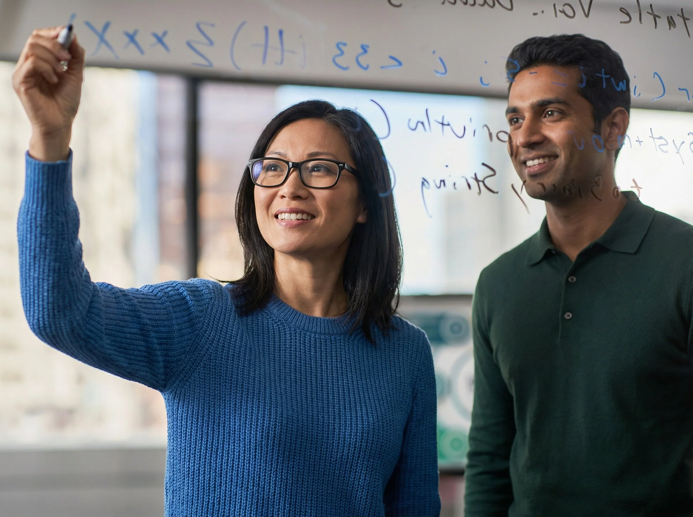A woman with black hair and glasses writing on a transparent board while a smiling man with dark hair looks on. The woman is wearing a blue sweater and the man a dark green polo shirt, in an office setting.
