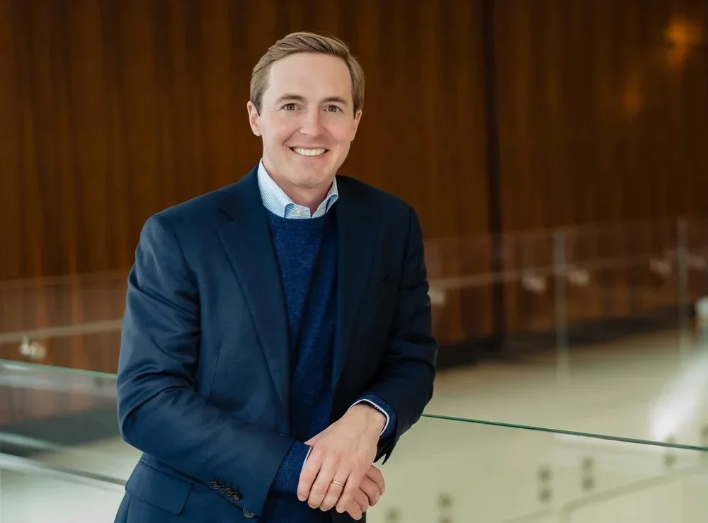 A smiling man in a blue suit standing indoors with a wooden background and glass railing.