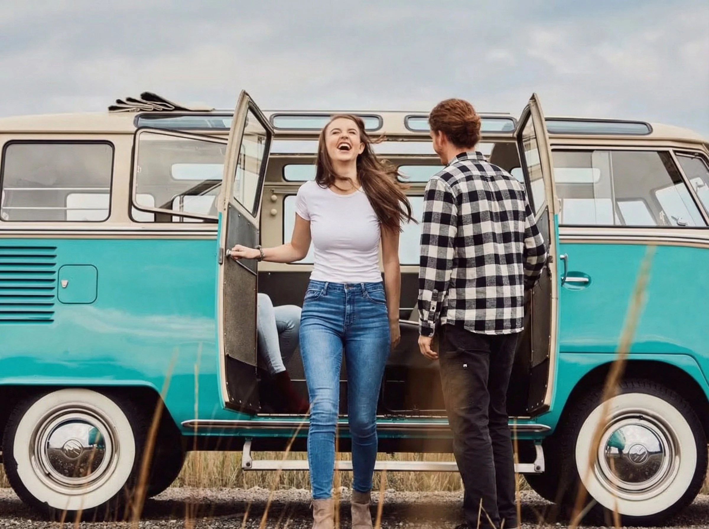 A woman and a man, both smiling and laughing, stand outside a vintage blue and white bus. The woman is holding the door open, and the man is facing her. The scene appears to be outdoors on a cloudy day.