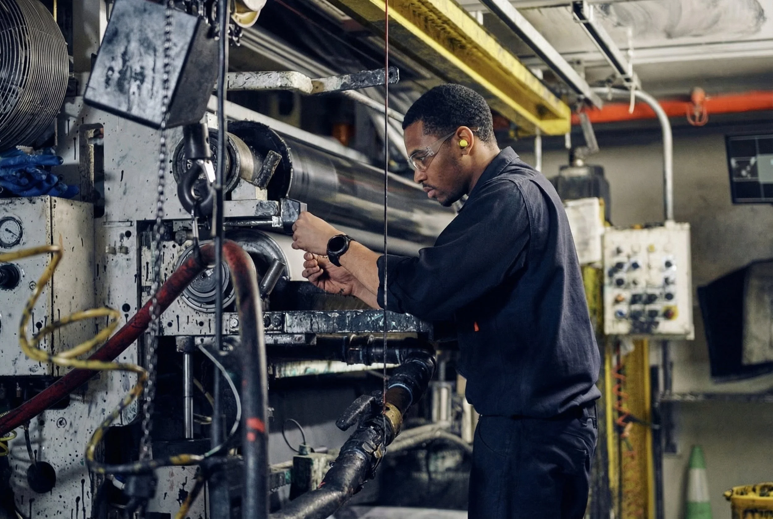 A man working on industrial machinery in a factory setting, wearing safety glasses, ear protection, a black uniform, and a watch.