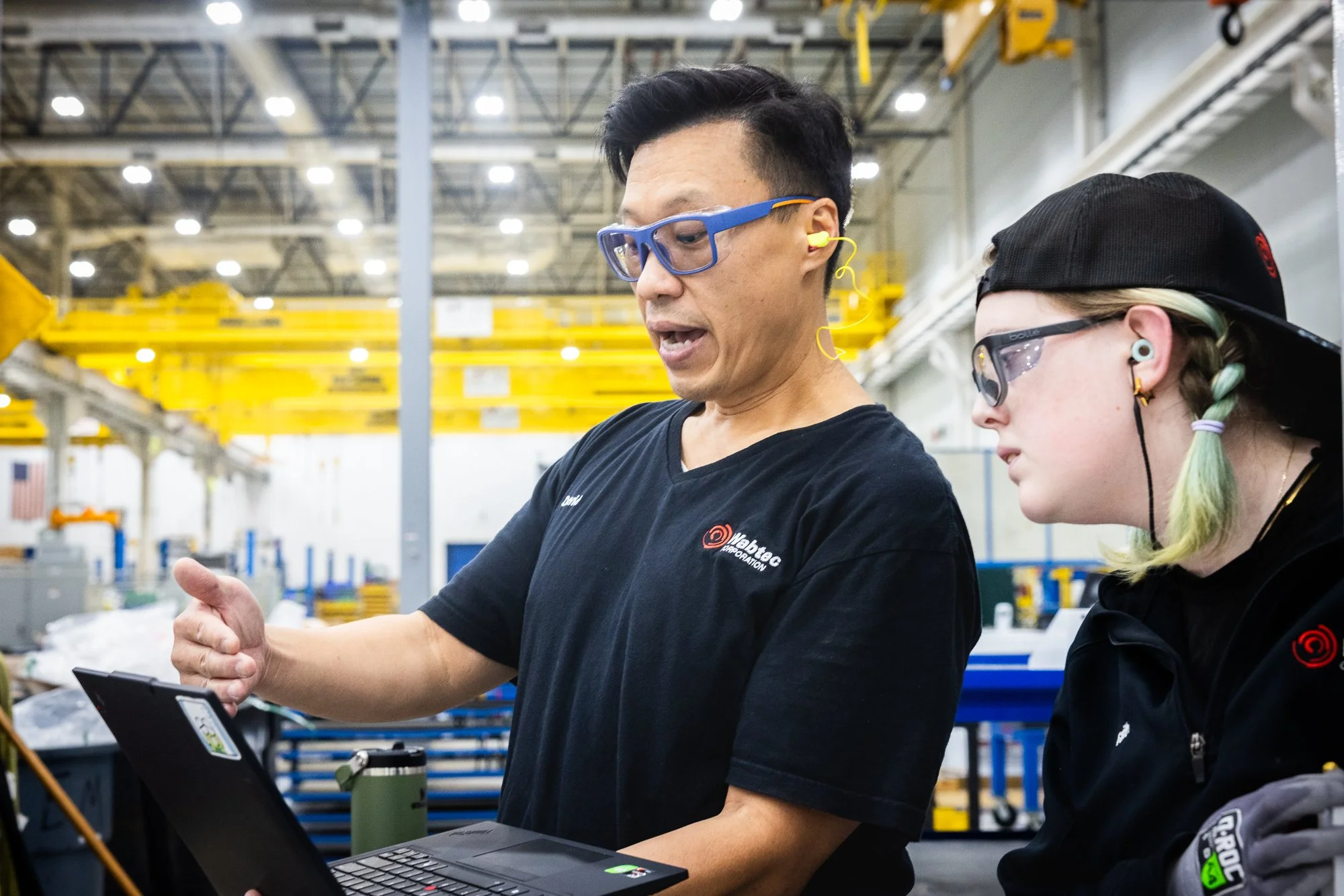 Two workers in a manufacturing or warehouse setting discussing information on a laptop, wearing safety glasses and ear protection.