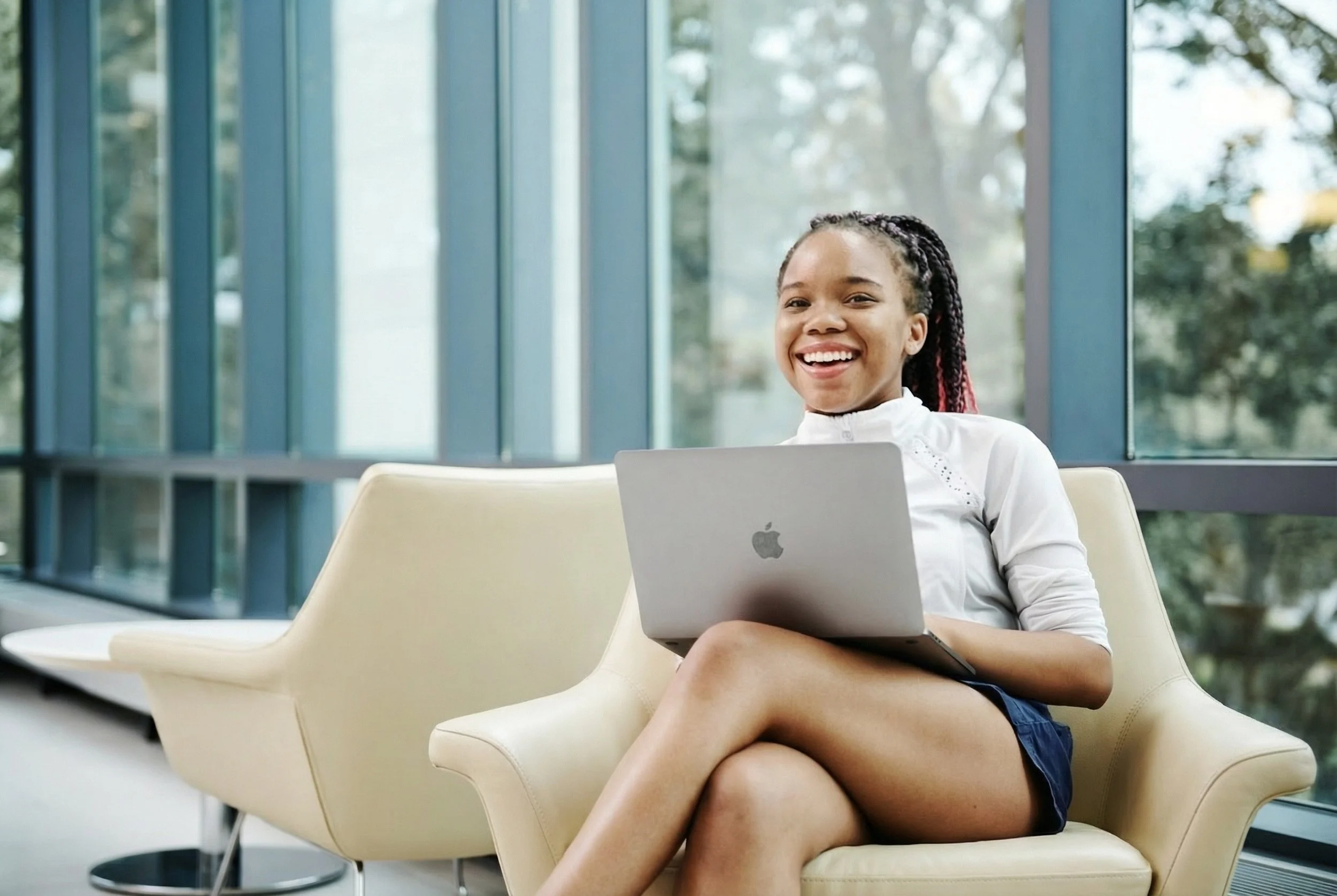 A young woman sitting on a cream-colored couch with crossed legs, smiling, holding a MacBook laptop in a modern, bright indoor space with large windows and greenery outside.