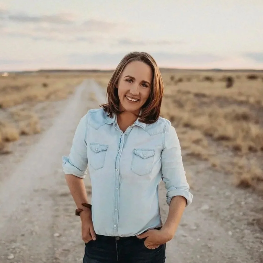 A woman with shoulder-length brown hair smiling and standing on a dirt road in a rural area, wearing a light blue denim shirt and dark pants, with arms relaxed and hands in her pockets.