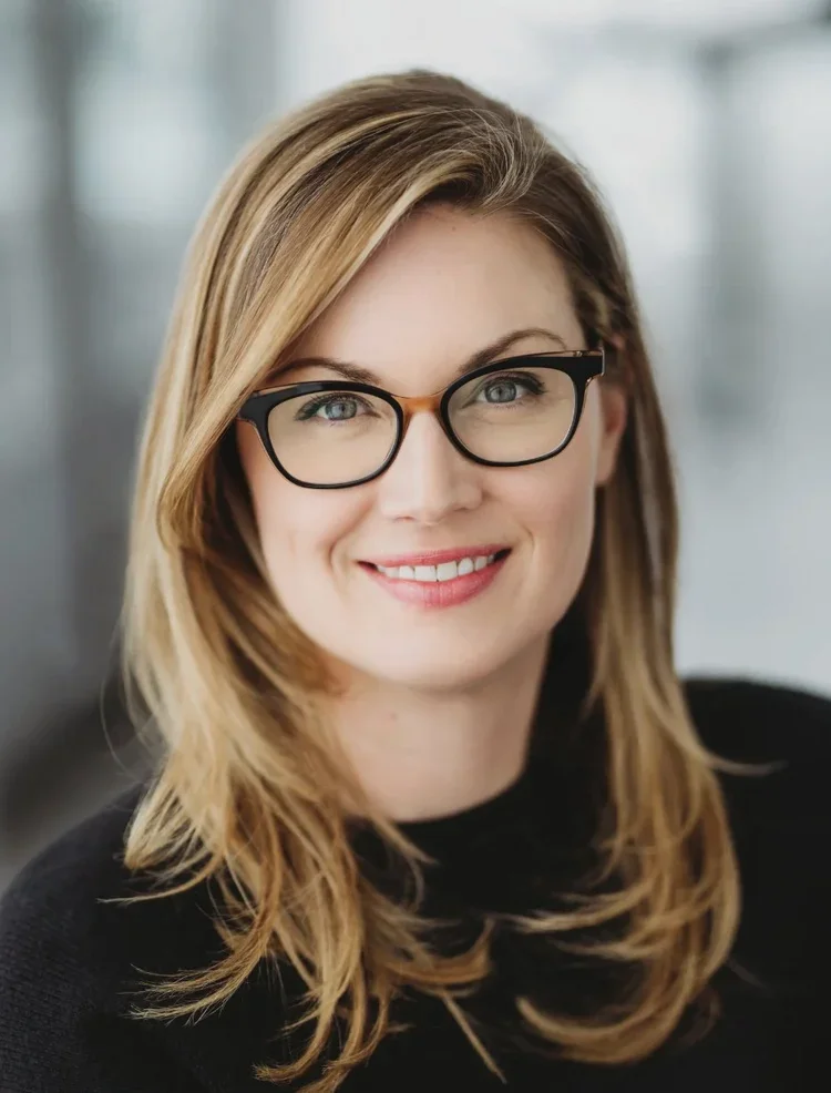 A woman with shoulder-length blonde hair, glasses, and a black sweater smiling indoors.