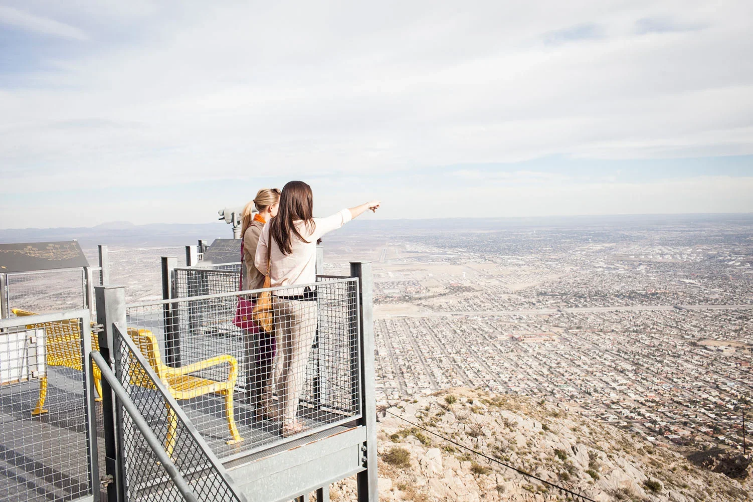 Three women standing on a viewing platform on a mountain, overlooking a cityscape below, with one woman pointing at something in the distance.