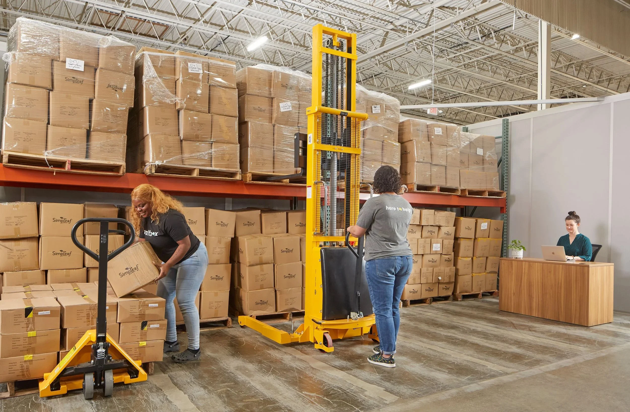 Warehouse workers moving boxes on pallets using a powered hand truck, with a woman working on a desk nearby.