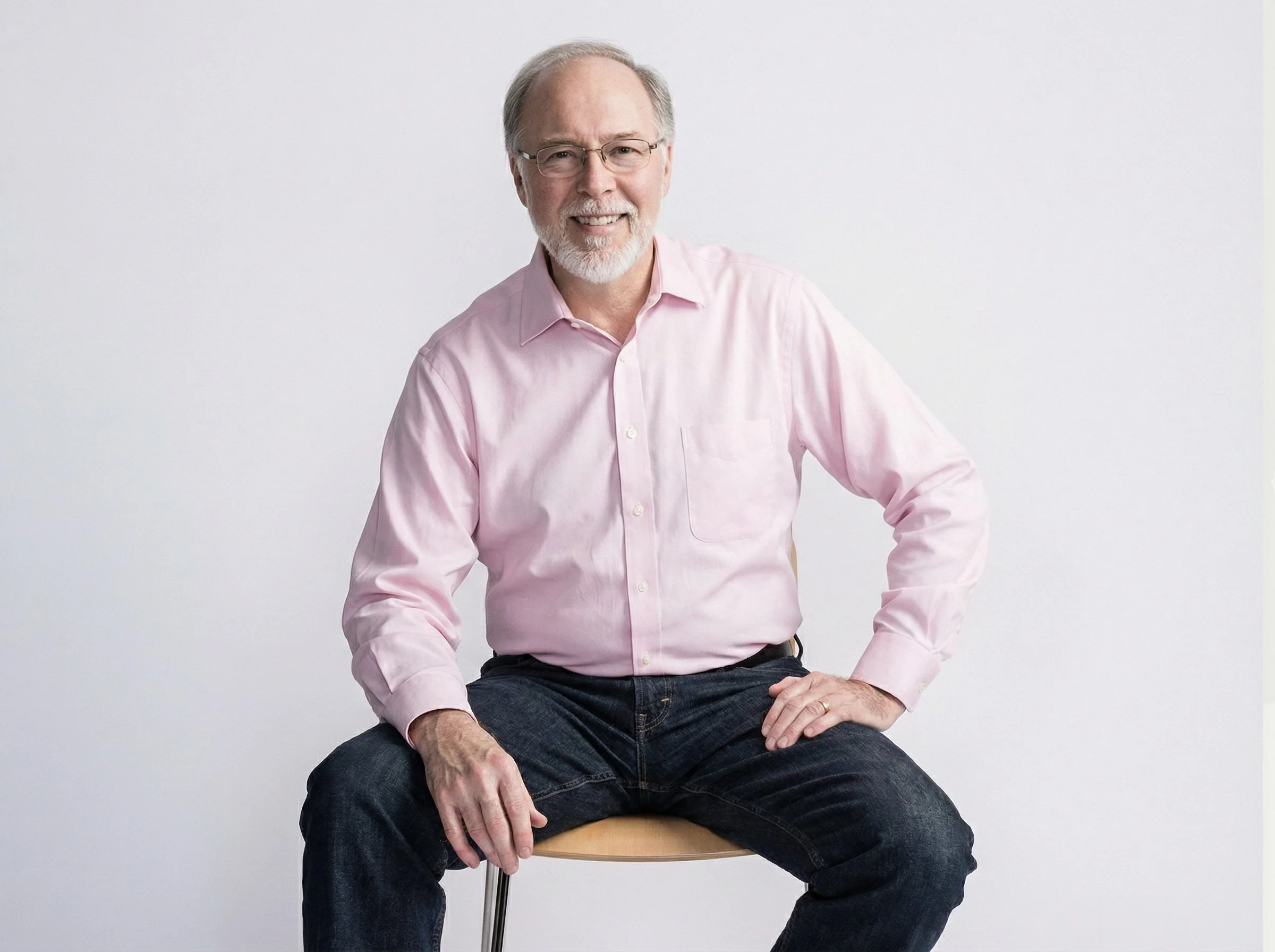 Older man with glasses and gray hair, wearing a pink button-up shirt and dark jeans, sitting on a stool against a plain white background.