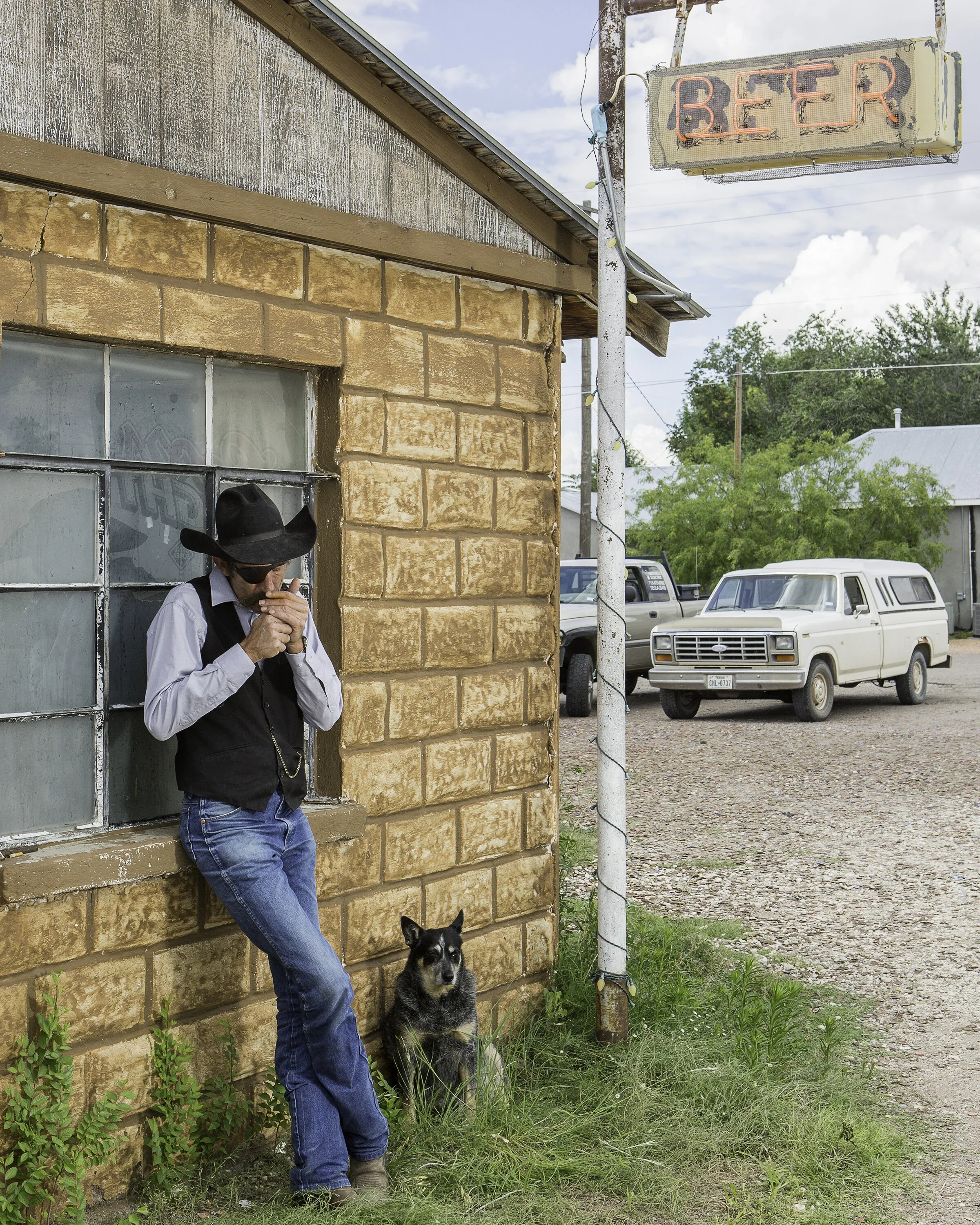 A man wearing a black hat, sunglasses, white shirt, and vest standing outside a brick building with a dog sitting beside him. There is a neon beer sign hanging from a pole and a gravel parking lot with vintage vehicles in the background.