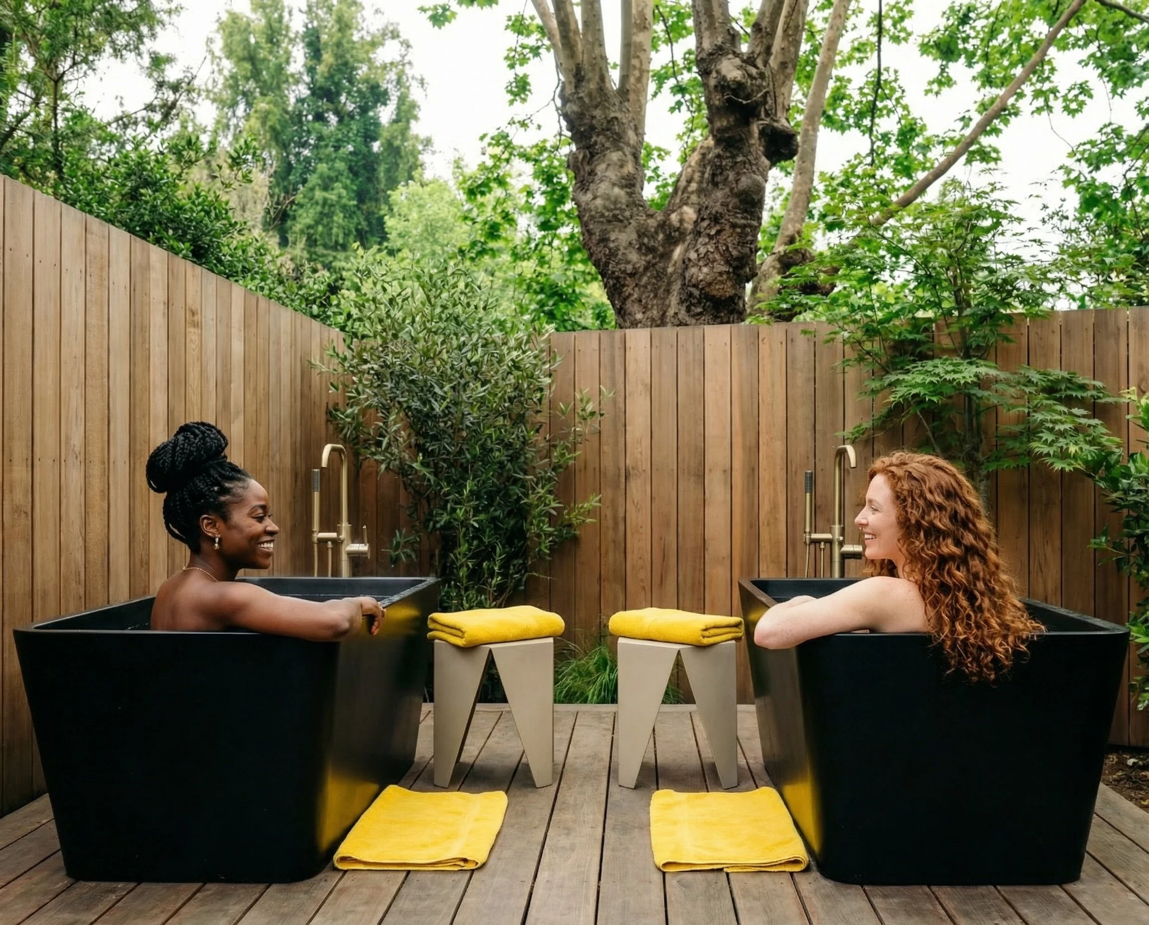 Two women relaxing in black bathtubs outdoors, facing each other and smiling, with yellow towels and small white stools between them, surrounded by a wooden fence, trees, and greenery.
