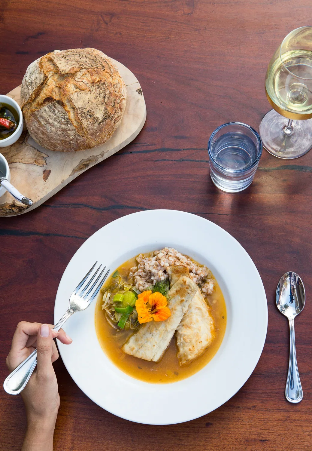 A plate with two pieces of fish, a scoop of chopped vegetables and grains, and a yellow flower garnish on a wooden table, with a glass of white wine, a glass of water, and a loaf of bread with olives nearby.