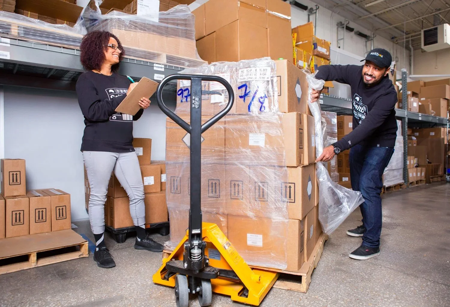 Two warehouse workers, a woman and a man, are handling large boxes on a pallet. The woman is smiling and holding a clipboard, while the man is lifting a box with a happy expression. They are in a storage facility with shelves filled with boxes.