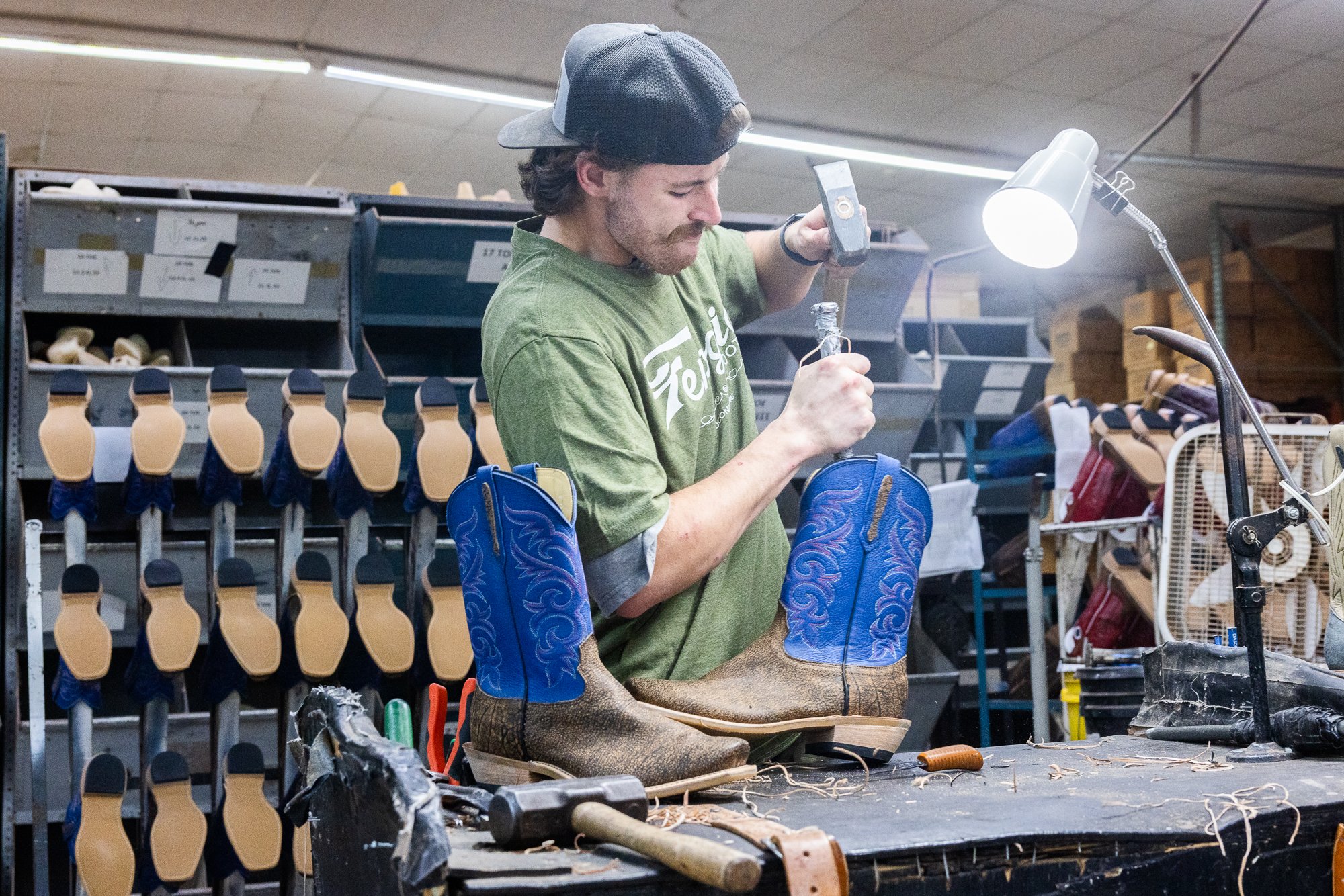 A man working on a pair of cowboy boots in a workshop. He is wearing a green T-shirt and a backwards baseball cap, using a tool on the boots which are blue with decorative stitching.