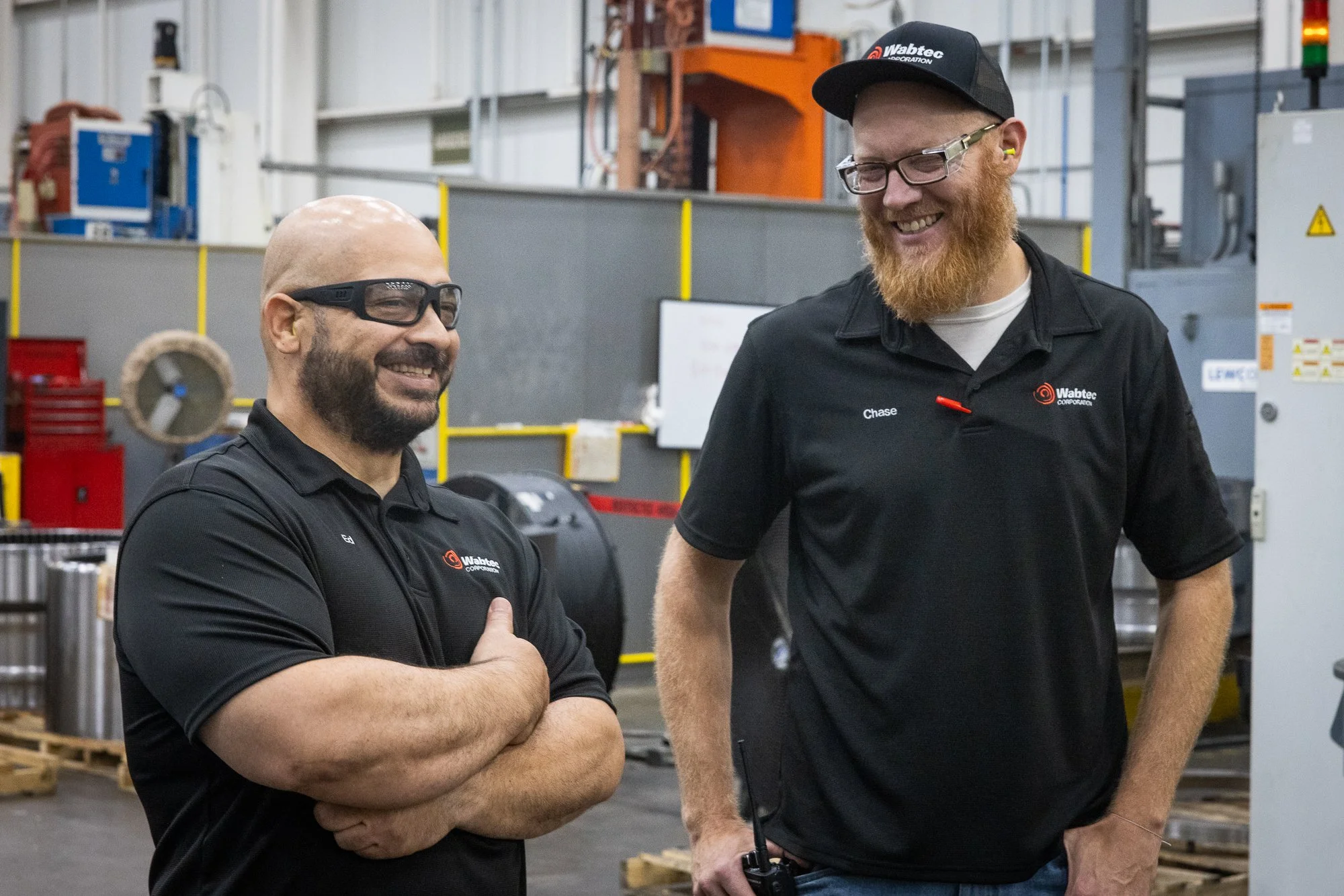 Two men in work uniforms smiling and having a conversation in an industrial setting, with machinery and equipment in the background.