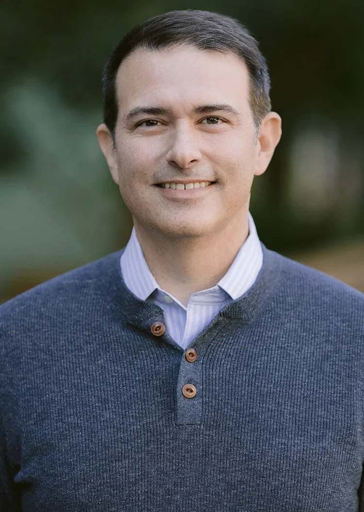 A man smiling outdoors, wearing a blue sweater over a collared shirt, with a blurred green background.