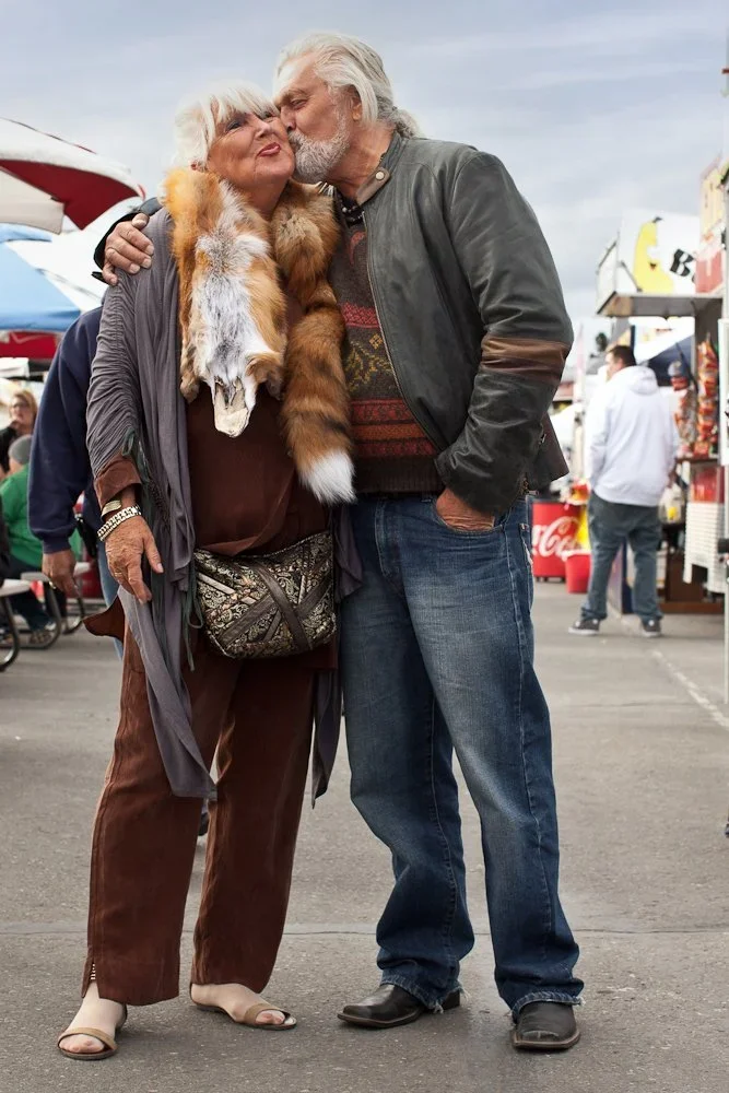 An elderly couple sharing a kiss at an outdoor market with tents, food stands, and other people in the background.