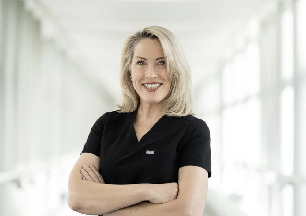 A woman with blonde hair smiling, wearing a black top, standing in a brightly lit, modern indoor space.