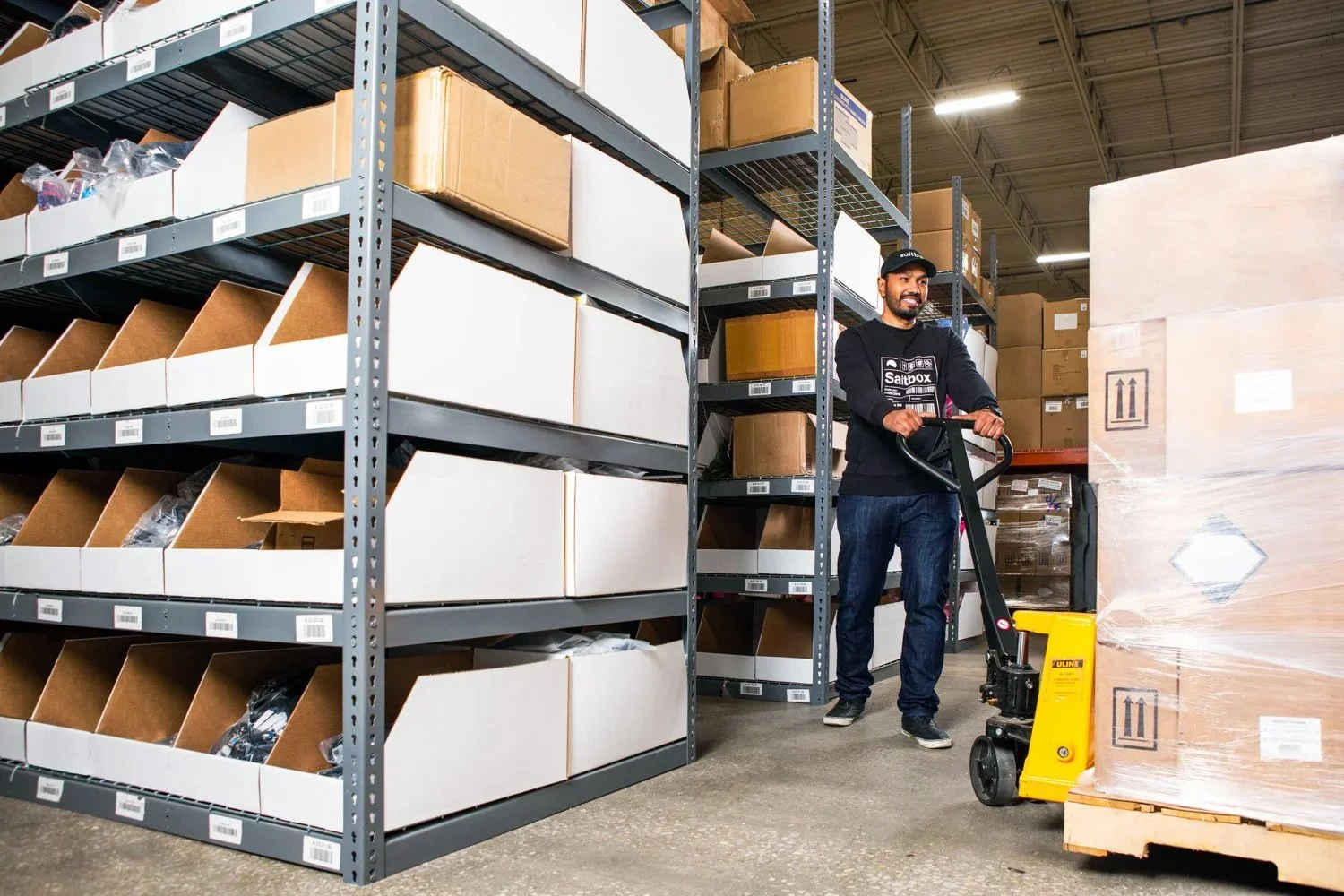 A man operating a yellow floor cleaner in a warehouse aisle, surrounded by metal shelves filled with boxes and supplies.