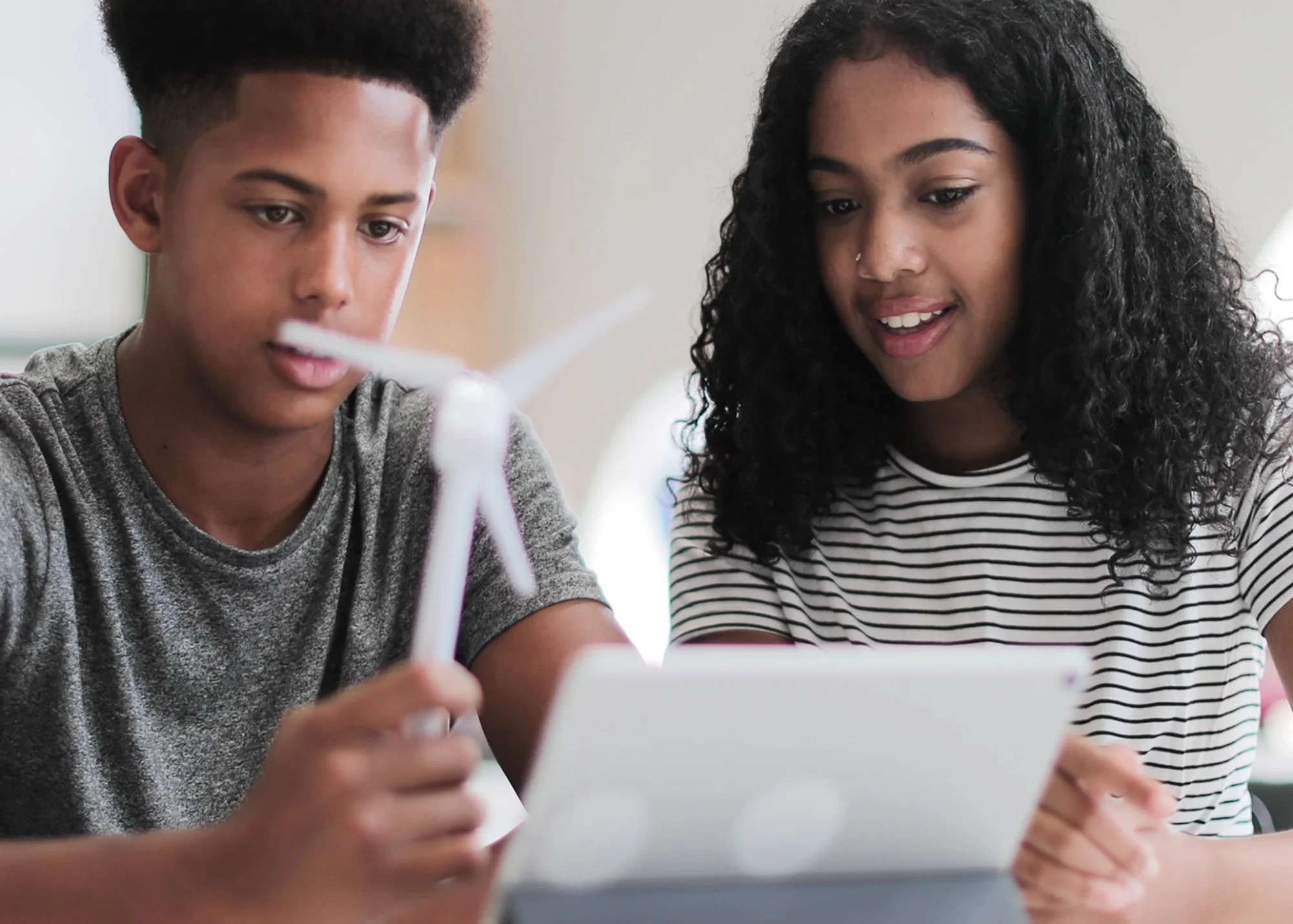 Two teenagers, a boy and a girl, sitting together and looking at a tablet, with the girl smiling.