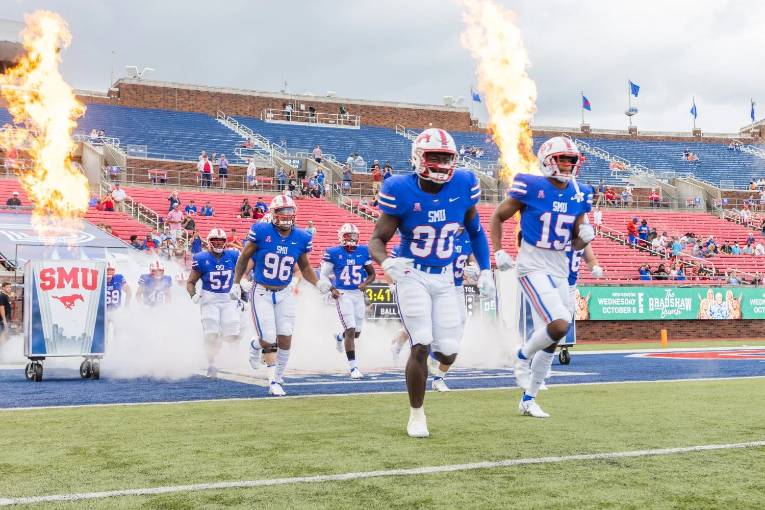 Football players from SMU team entering field with flames and smoke effects, fans in stadium watching.