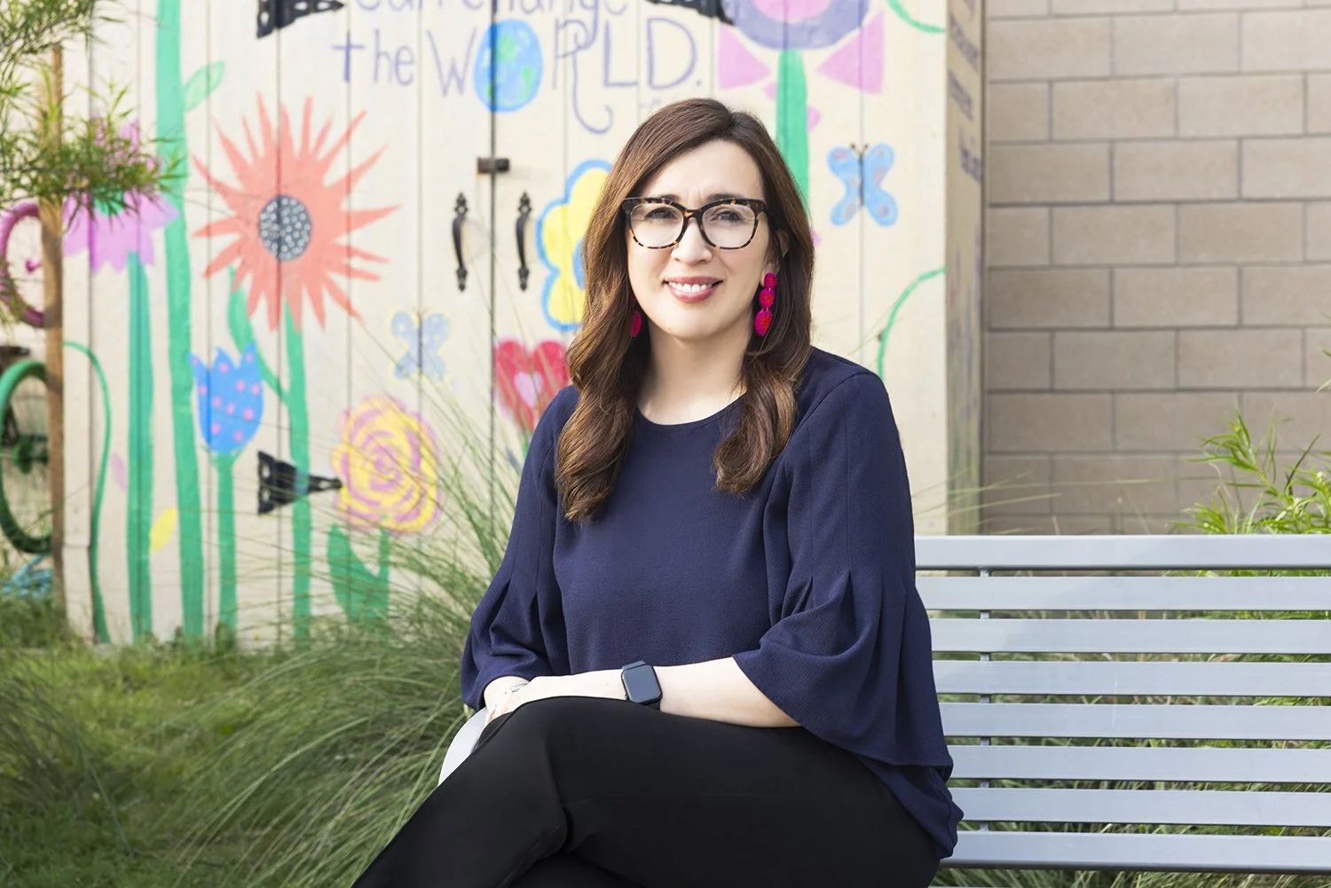 A woman with glasses and earrings sitting on a bench in front of a colorful mural with flowers on a wall.