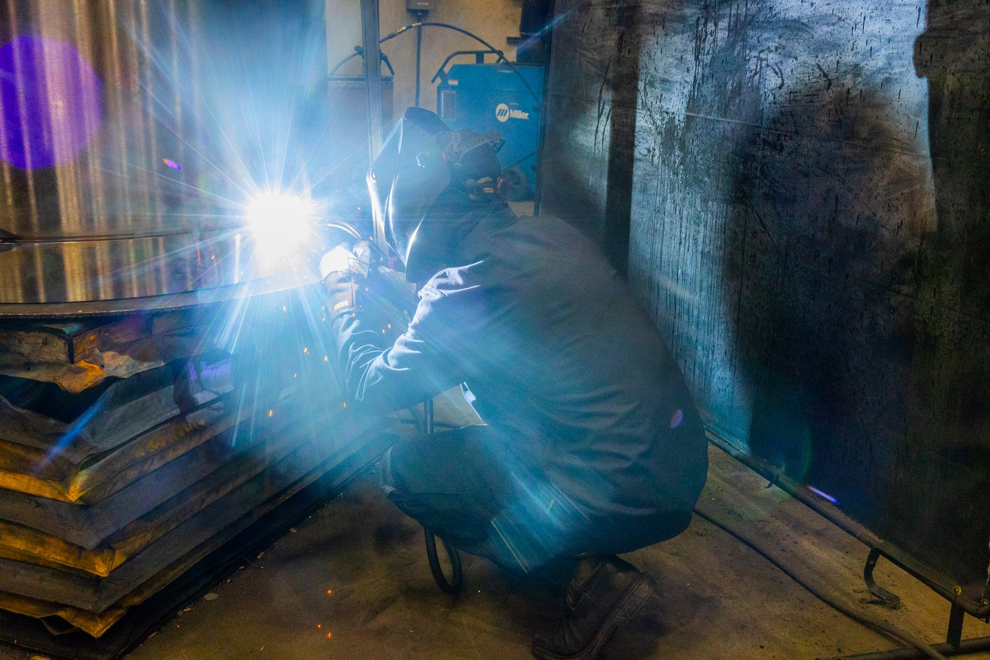 A person welding metal in an industrial workshop, wearing protective gear and helmet, with bright sparks from the welding process.