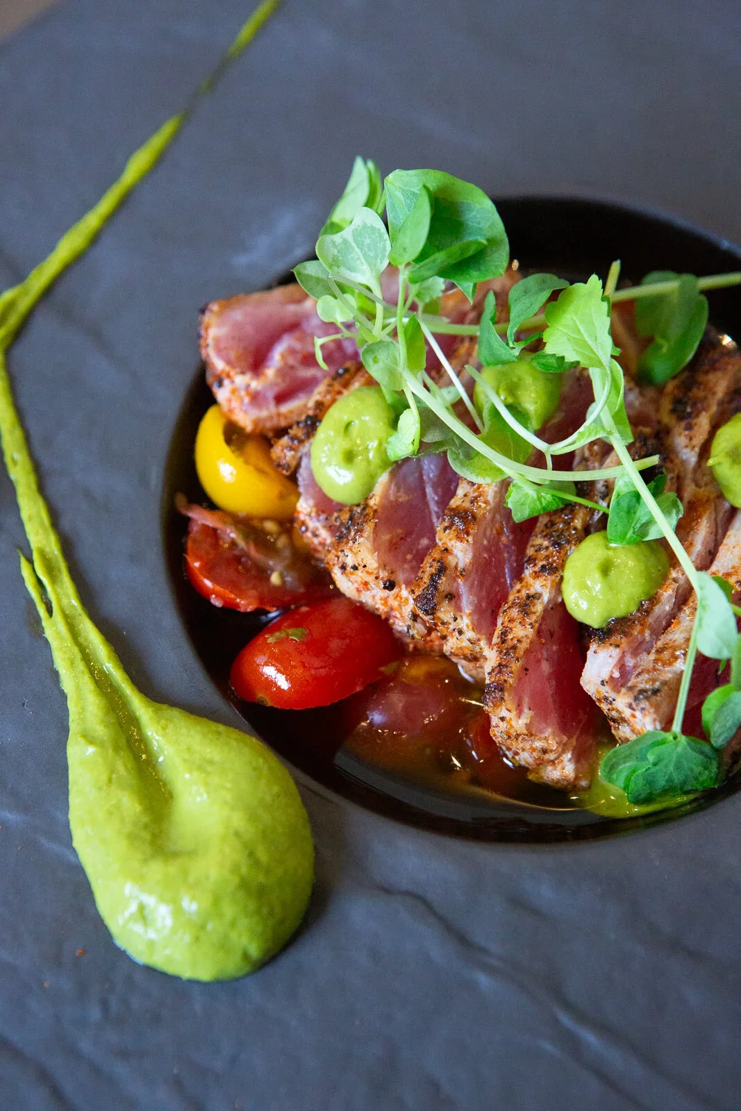 Seared tuna steak with microgreens, cherry tomatoes, and green sauce on a black plate with a green smear on dark slate background.