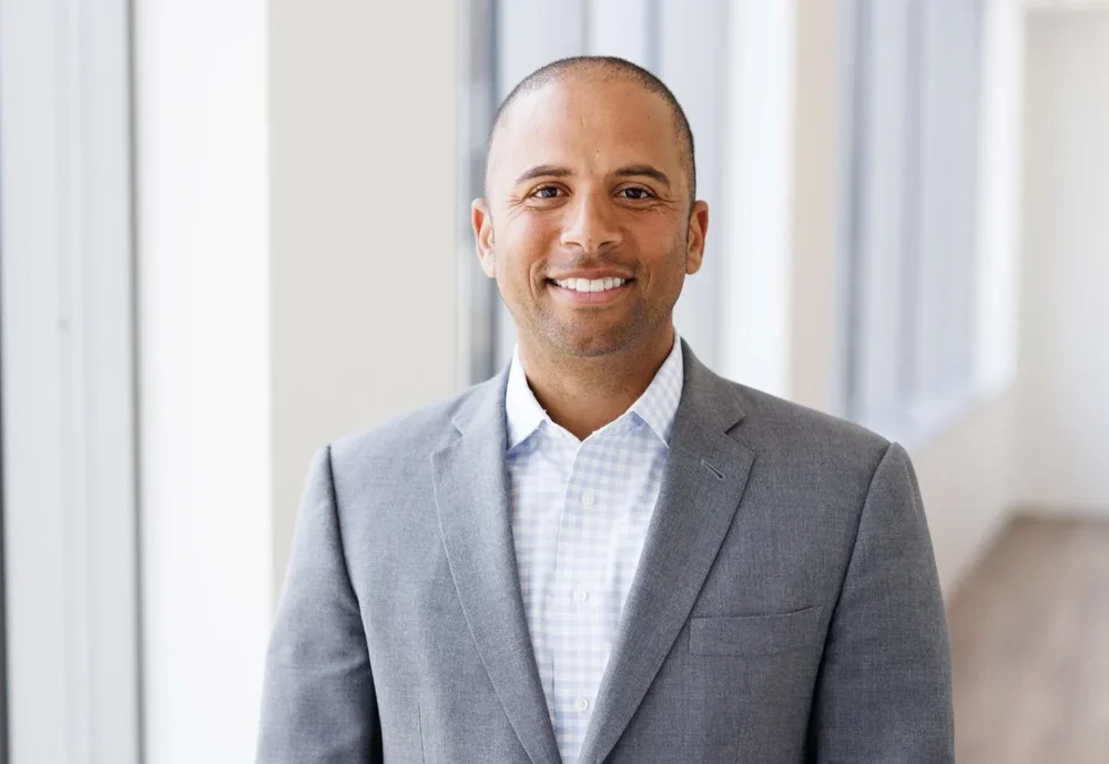 A man in a gray suit and white shirt standing in a bright office corridor, smiling at the camera.