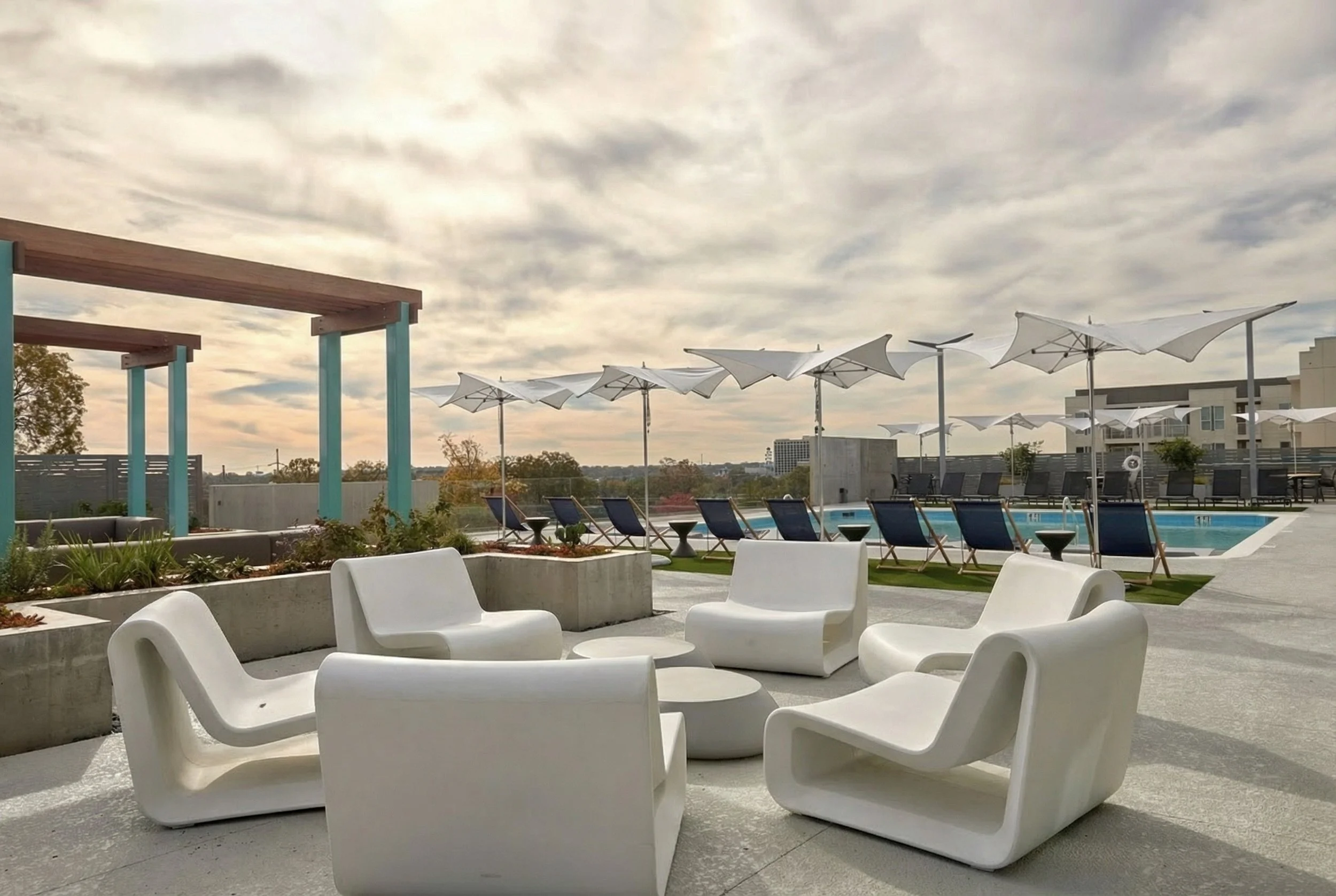 Rooftop outdoor pool area with white modern chairs in the foreground, poolside lounge chairs with umbrellas in the background, and a cloudy sky overhead.