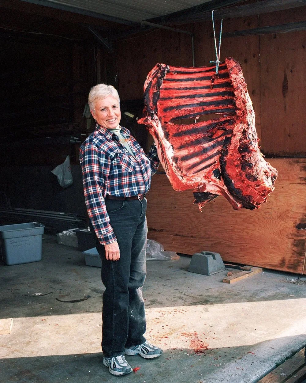 A smiling woman in a plaid shirt standing next to a large hanging slab of meat with ribs, inside a butcher's shop or meat processing area.