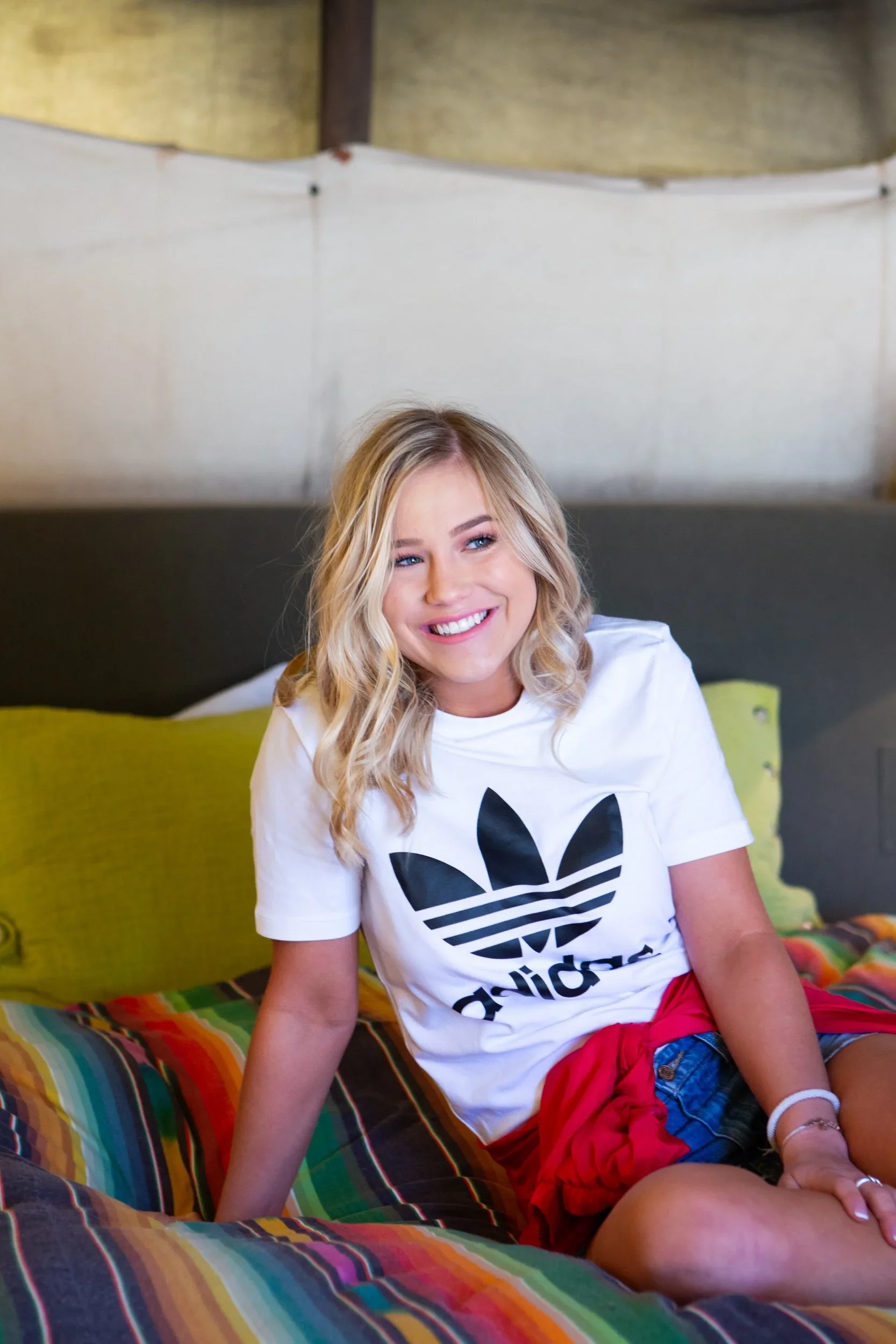 A young woman with blonde hair sitting on a bed, smiling, wearing a white Adidas t-shirt and red shorts, with colorful striped bedding.