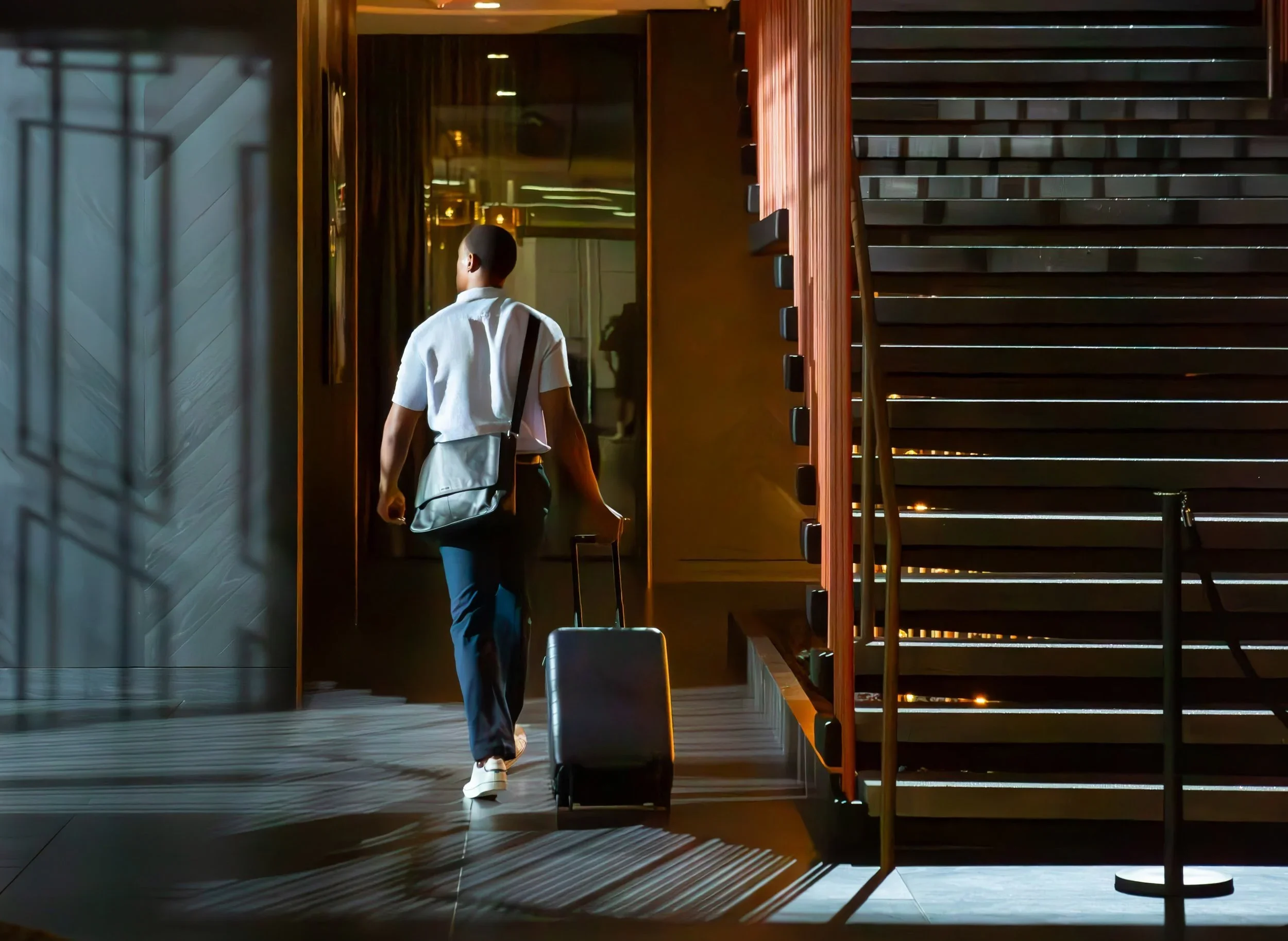 A man walks through a hotel lobby carrying a suitcase and a shoulder bag, with sunlight casting shadows on the floor.