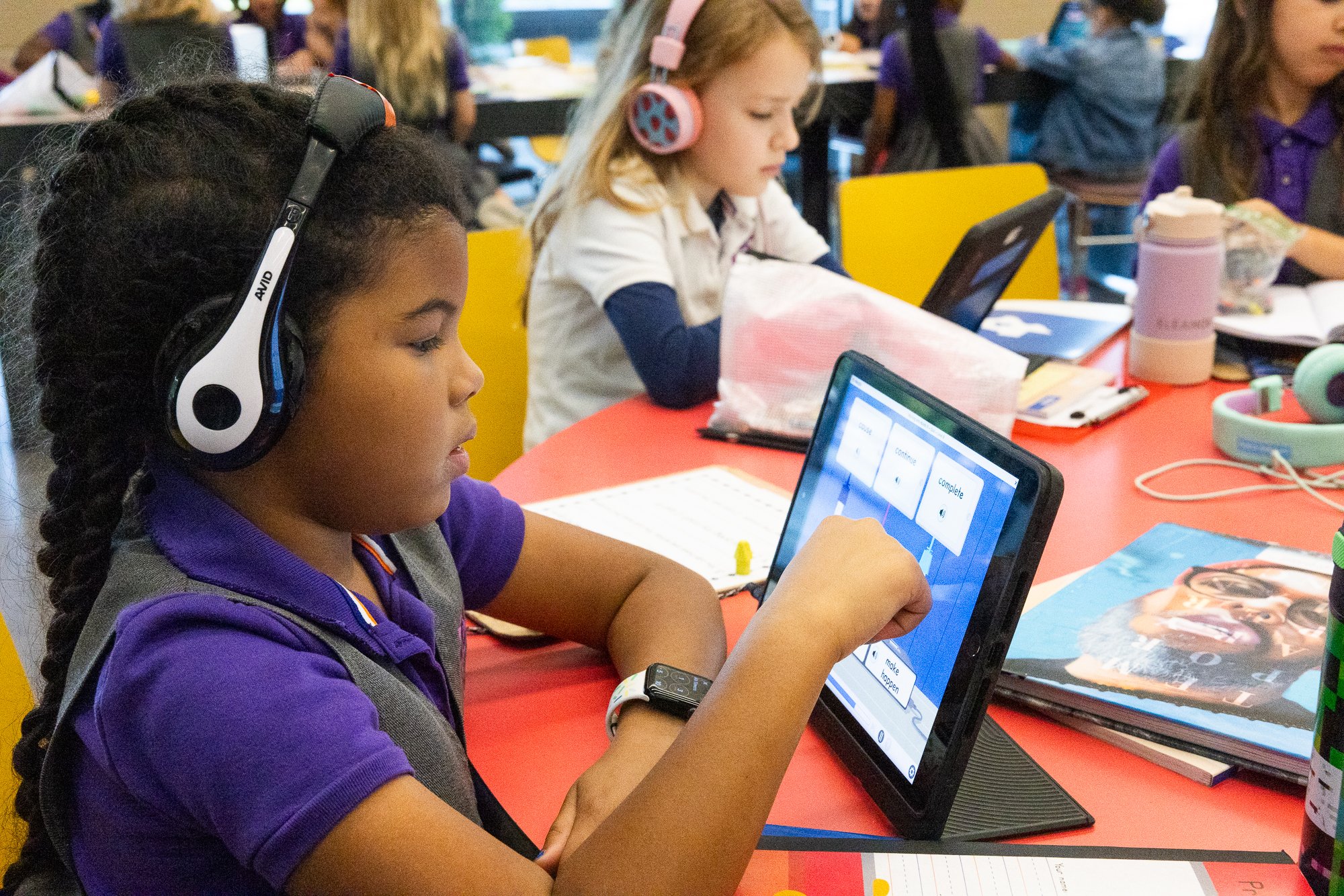 A young girl using a tablet, wearing headphones, in a classroom with other children also working, some with tablets or notebooks on the table, colorful chairs, and various school supplies.