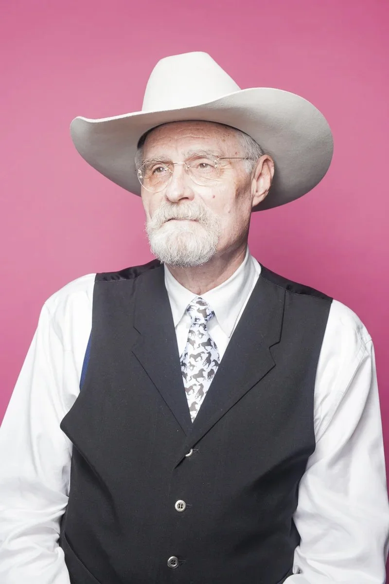 An elderly man with a white beard and glasses wearing a white cowboy hat, a black vest over a white shirt, and a patterned tie against a pink background.