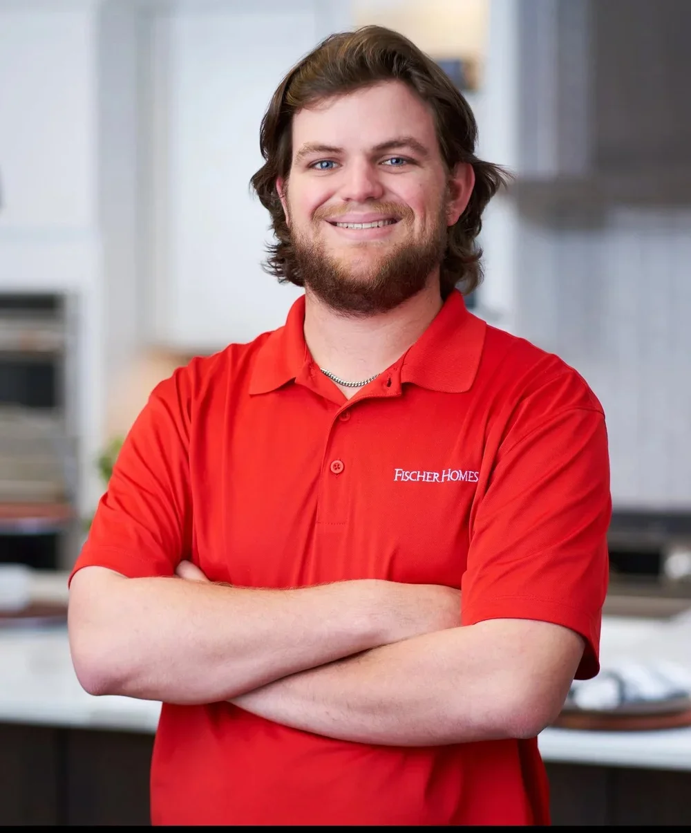 A man with brown hair and a beard smiling, wearing a red polo shirt with 'FISCHER HOMES' embroidered on it, standing with arms crossed in an indoor setting.