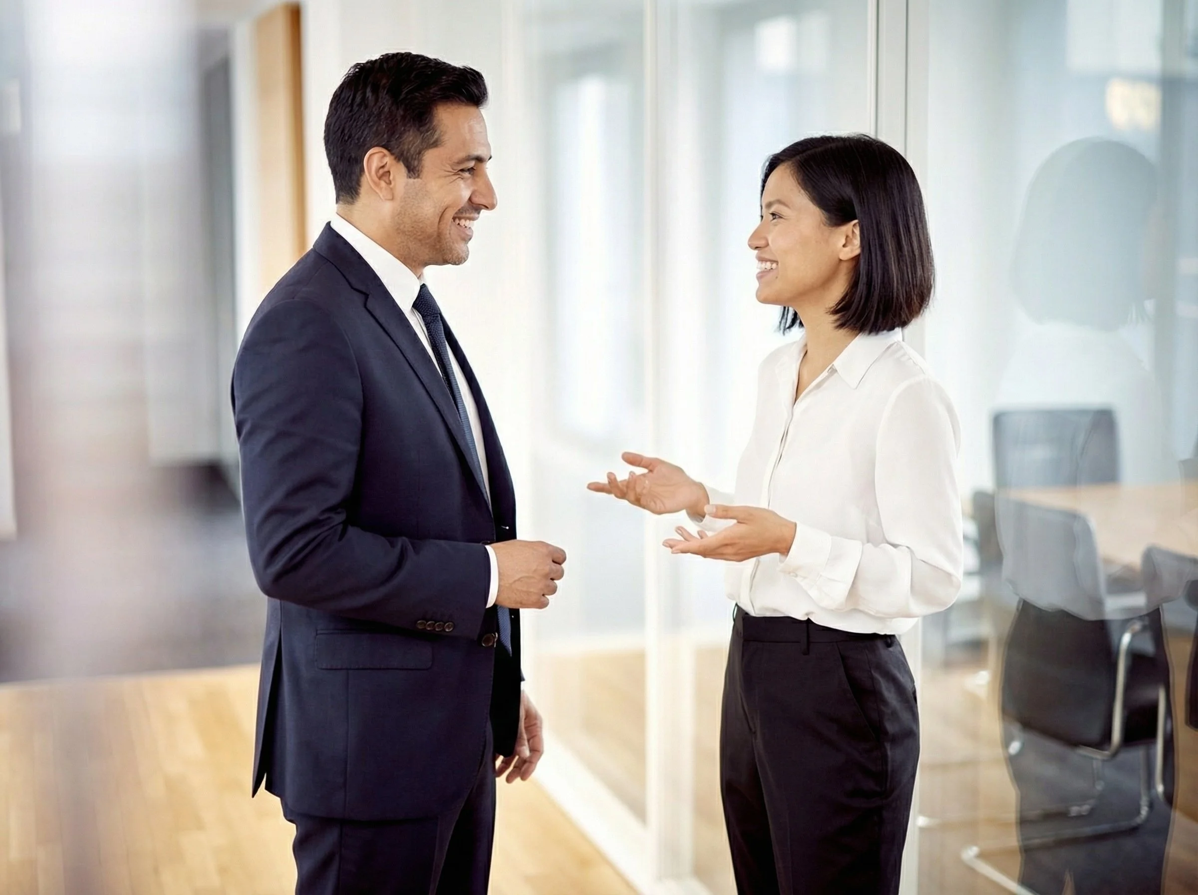 A man in a navy suit and a woman in a white shirt having a conversation in an office.