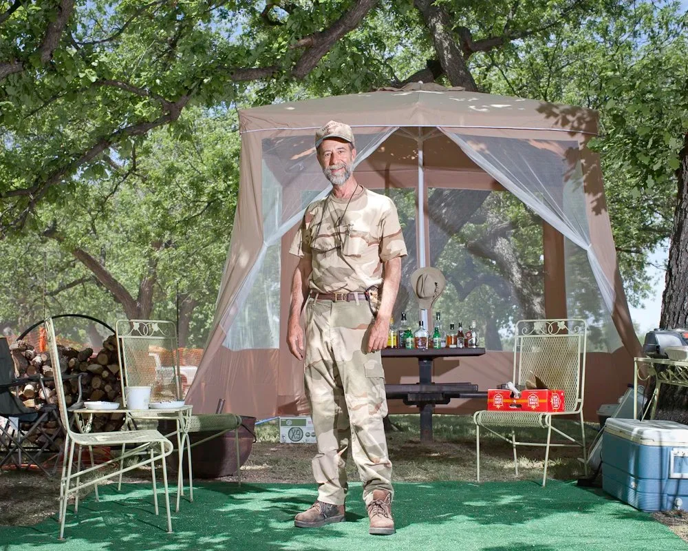 A man in camouflage military uniform standing outdoors behind a camping shelter, with trees in the background and outdoor furniture surrounding him.