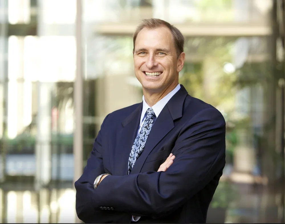 A smiling man in a dark suit and tie standing outdoors with arms crossed, in front of a modern building with glass windows.