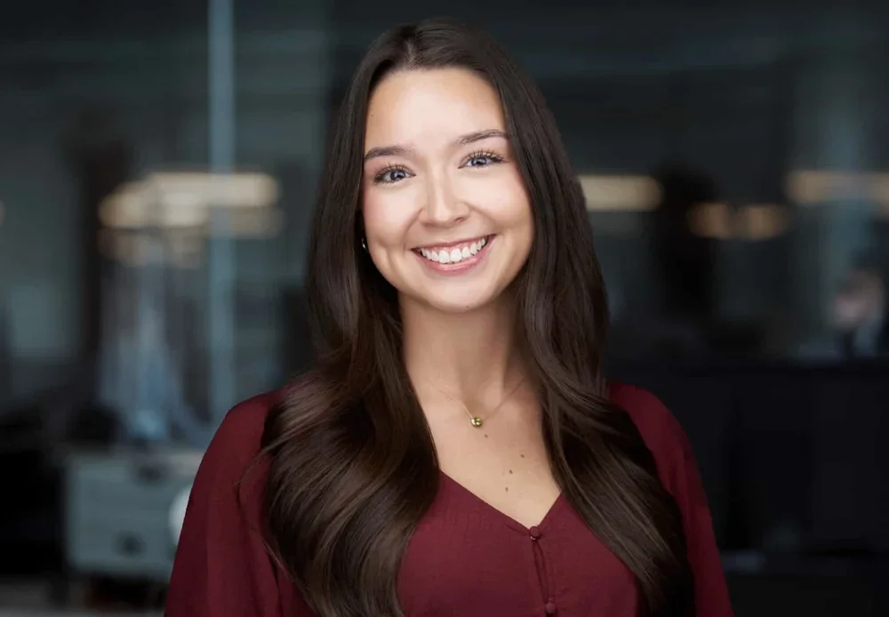 A young woman with long, wavy brown hair, smiling and wearing a burgundy top, standing in a modern office setting.